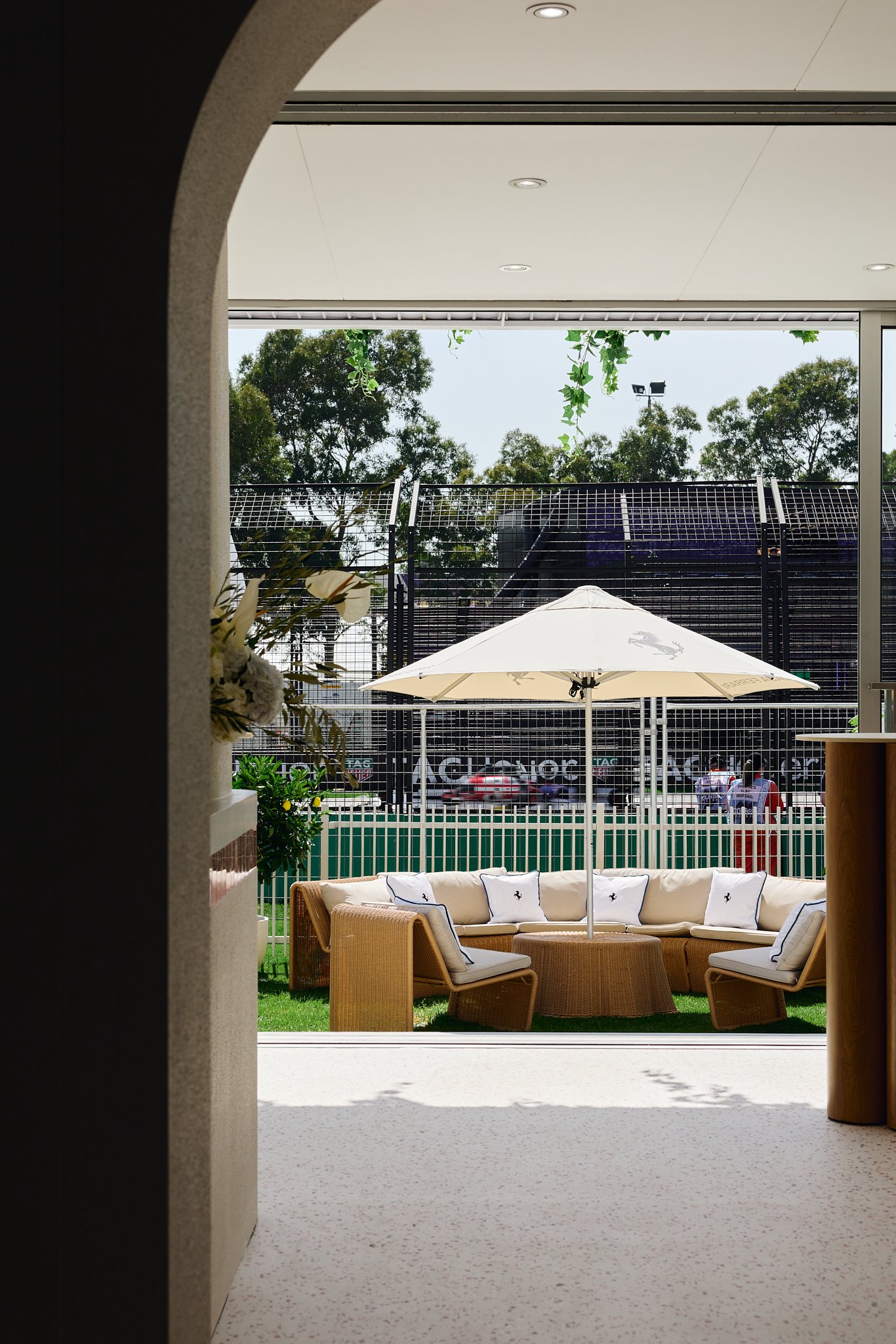 View through a doorway of an outdoor seating area with a beige curved sectional sofa, white cushions with dark piping, a round coffee table, a large white umbrella, a grassy lawn, and tall trees in the background.