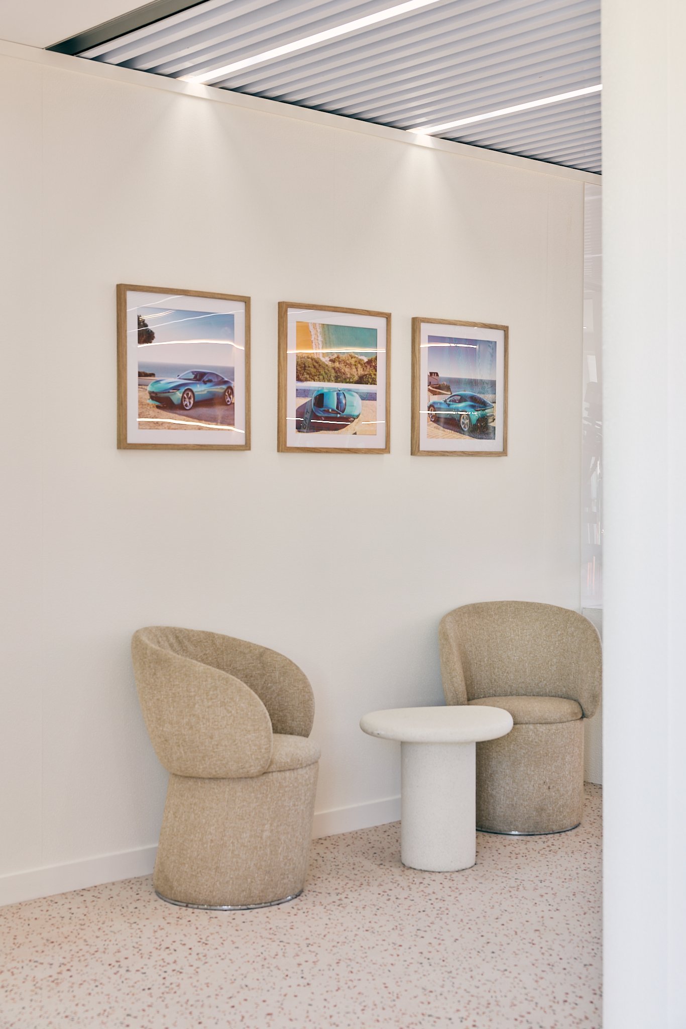 Interior of a modern waiting area with two beige chairs and a small white round table between them. Three framed pictures of sports cars and scenic landscapes hang on a white wall. The ceiling has a metallic design with linear lighting.
