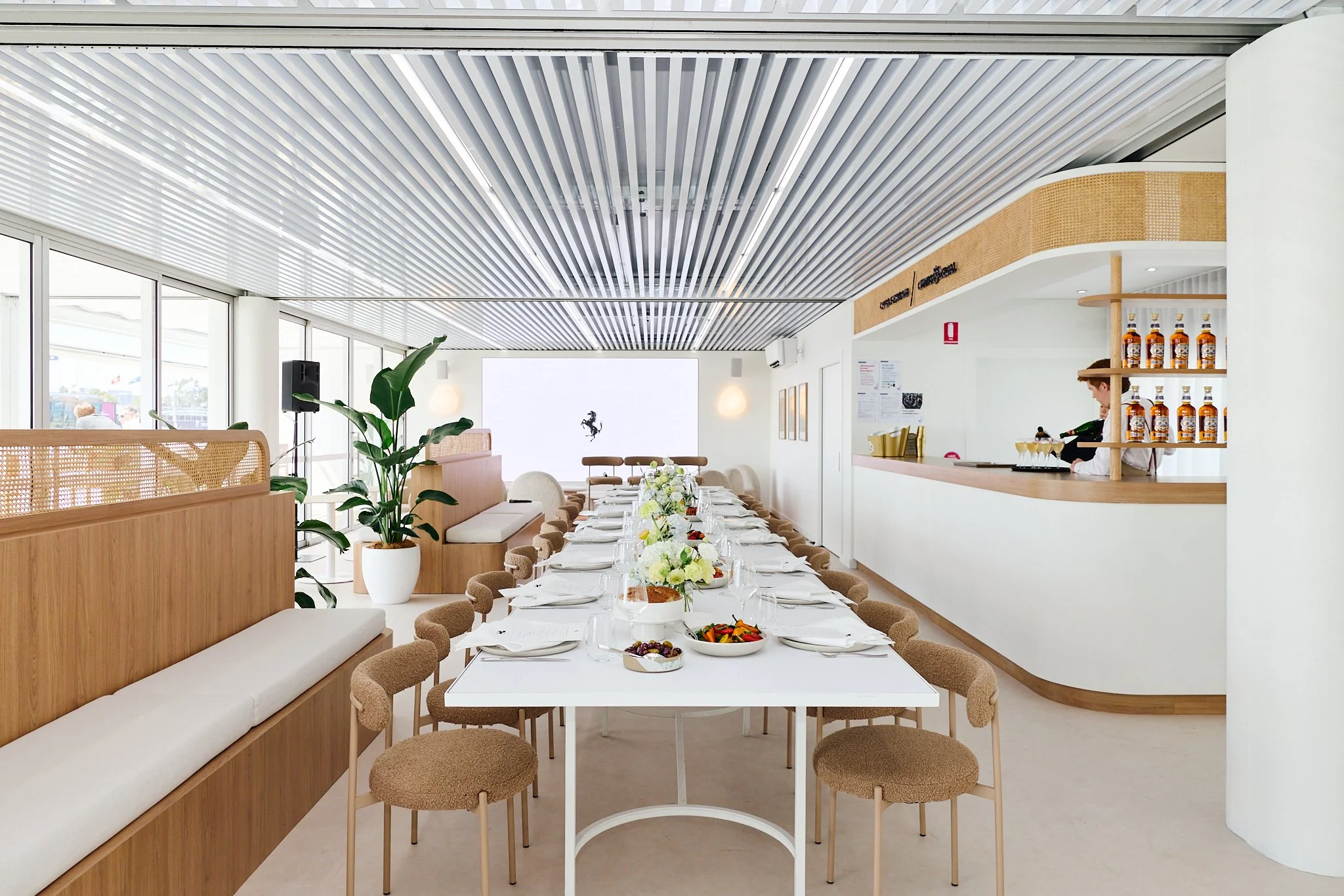 A modern restaurant with a long dining table set with plates, glasses, and bowls, surrounded by beige chairs and banquettes. The room features large windows, decorative plants, a white ceiling with striped panels, and a bar area with a person prepari