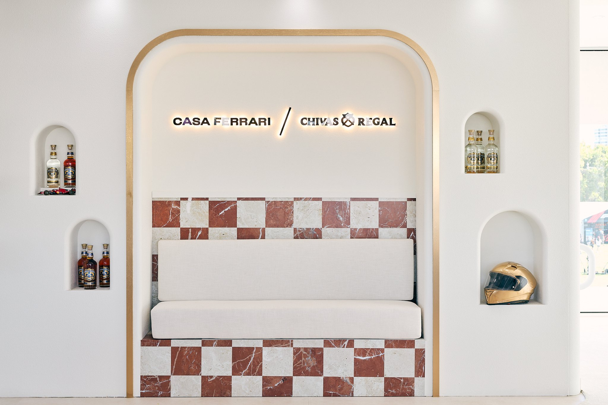 Modern reception area with white bench, red and white marble tiles, sign with logos of Casa Ferrari and Chivas Regal, and bottles of liquor on built-in shelves.