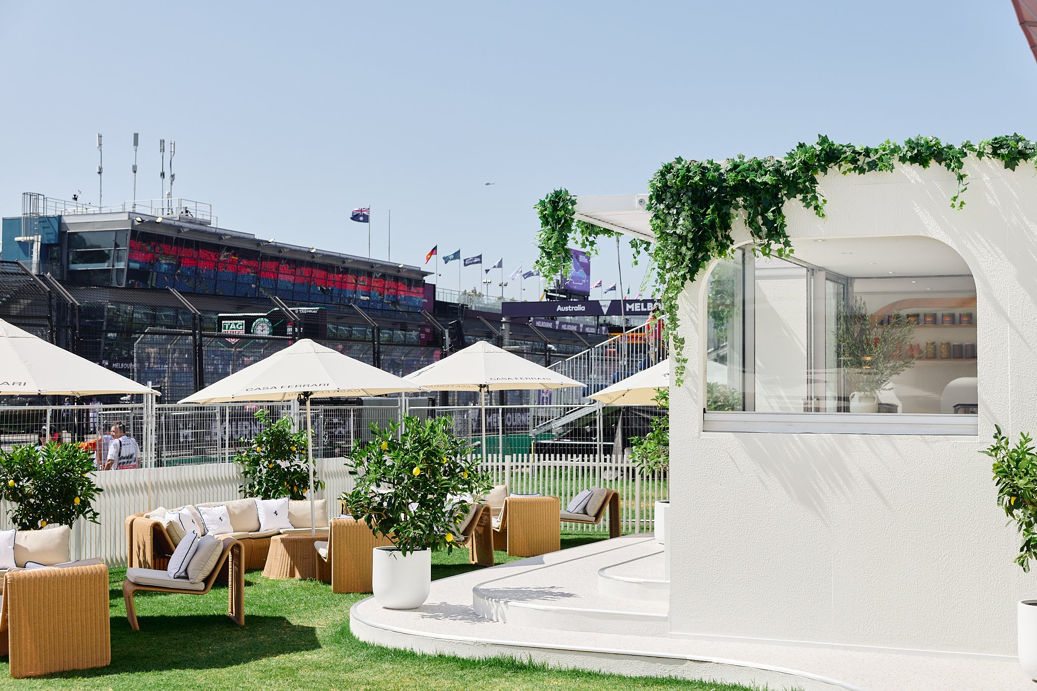 Outdoor area of a luxury sports event venue with white furniture, umbrellas, potted plants, and a view of the race track and grandstands.