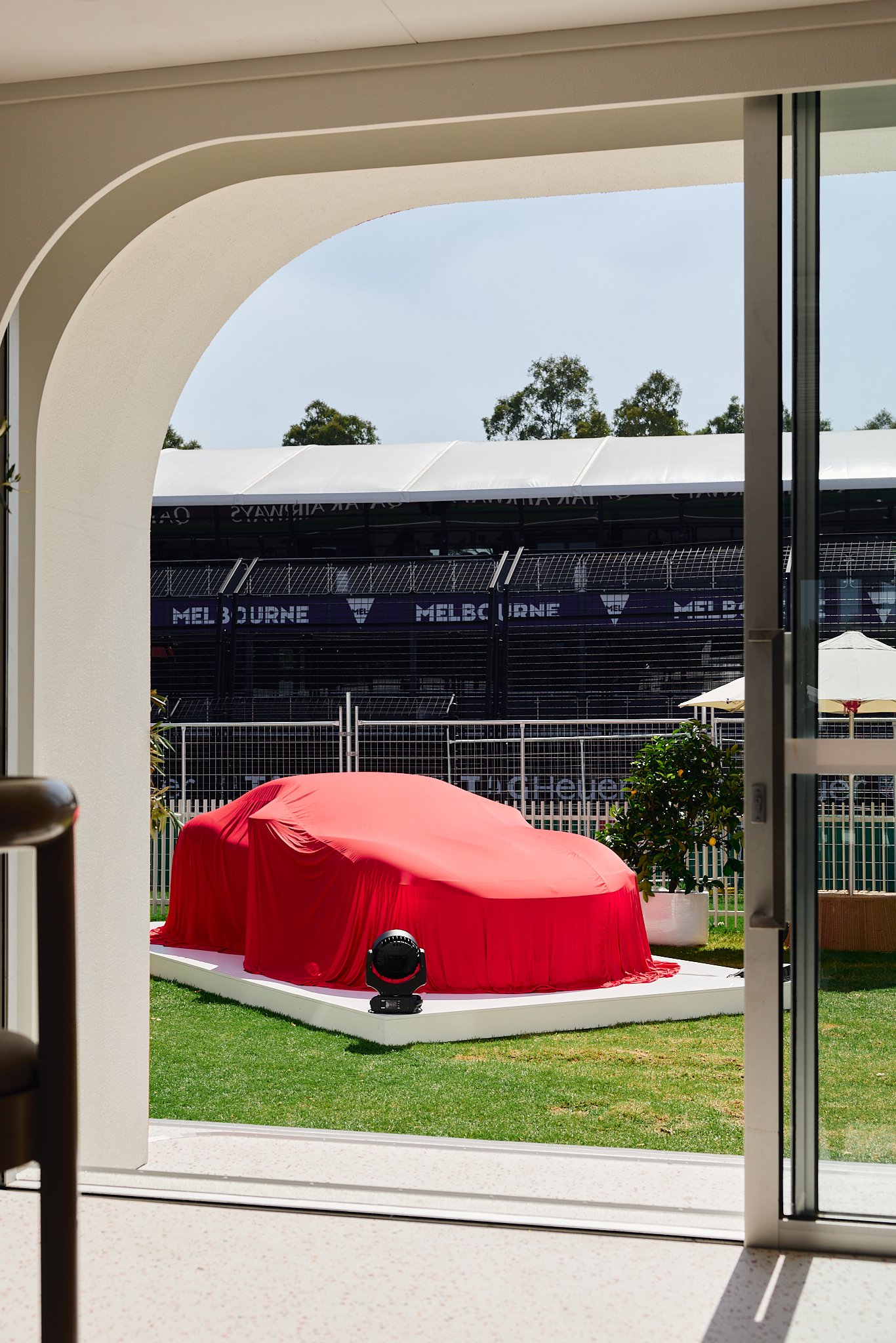 Looking through a doorway, a covered sports car on a white platform outside. The car is draped with a red cloth, with a black spotlight illuminating it. In the background, a racetrack with 'Melbourne' signage and fencing is visible.