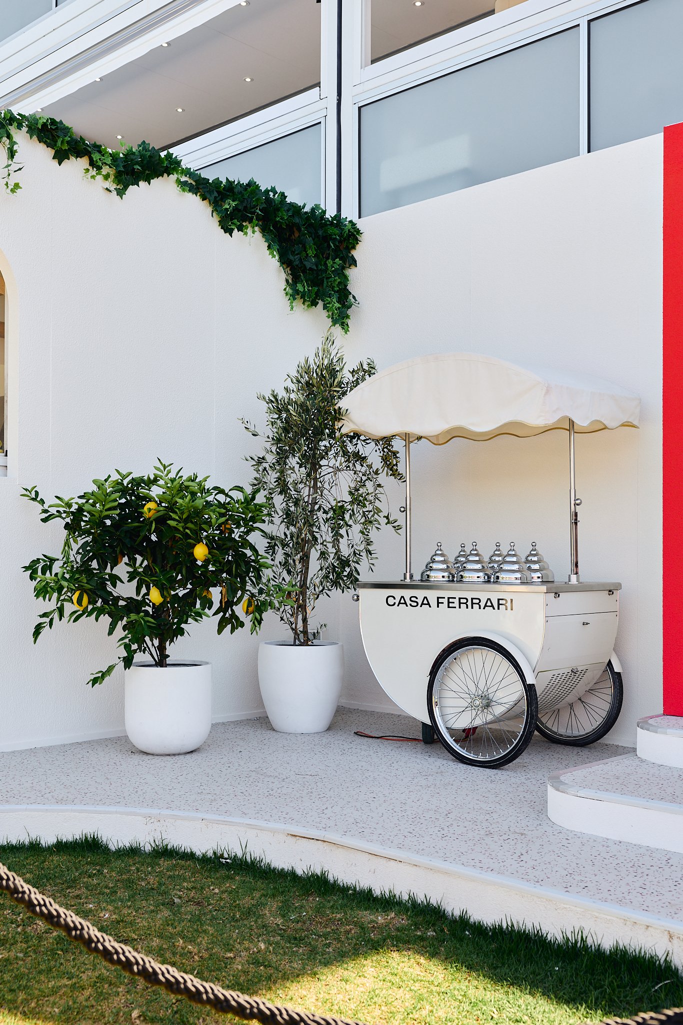 A mobile ice cream or gelato cart with 'Casa Ferrari' written on it, set on a white terrace with potted lemon trees and greenery, against a white wall and modern building with glass windows.