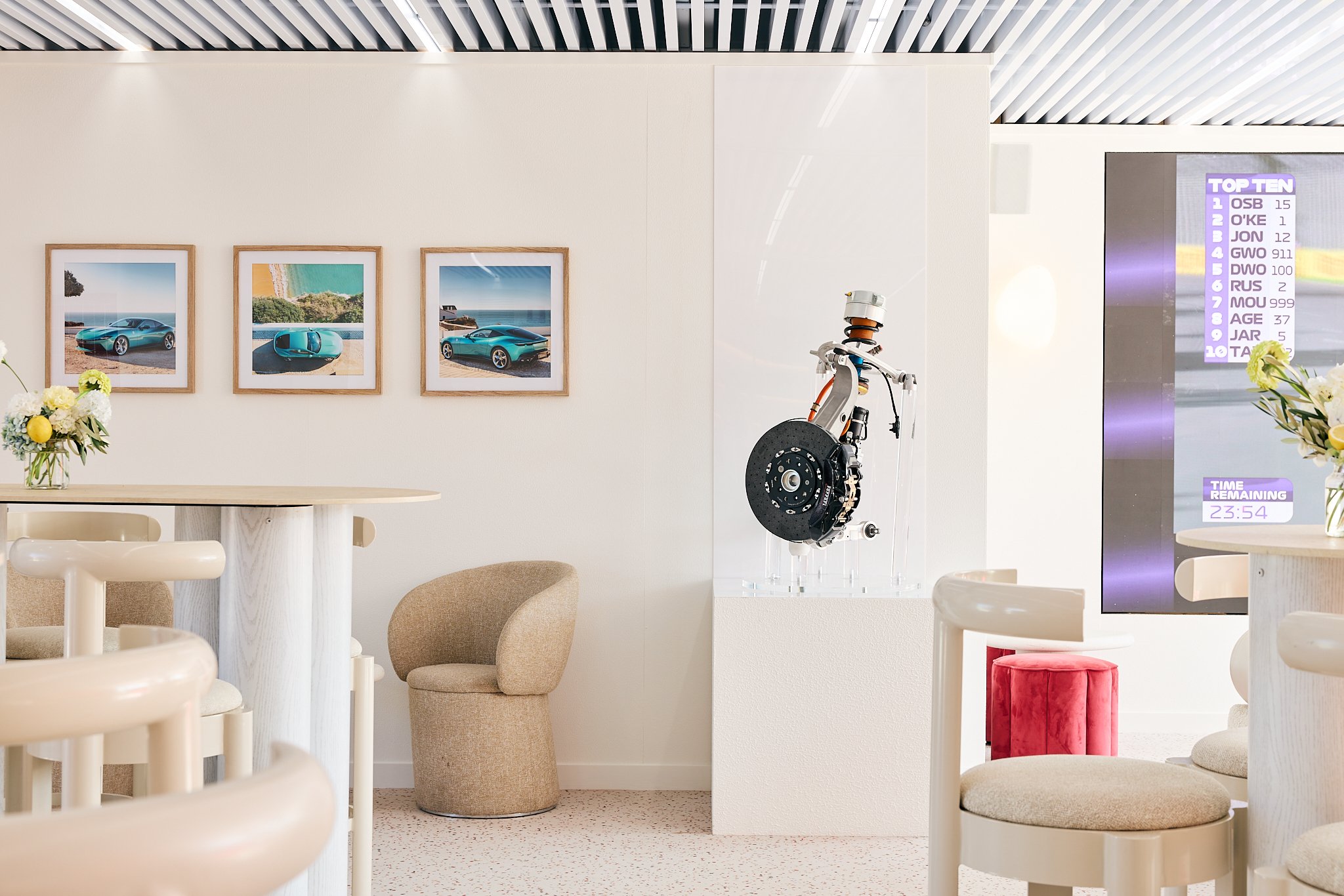 Modern interior with framed car photos on wall, beige chairs, a round table with a flower vase, a suspended car engine on display, and a digital scoreboard showing race results.