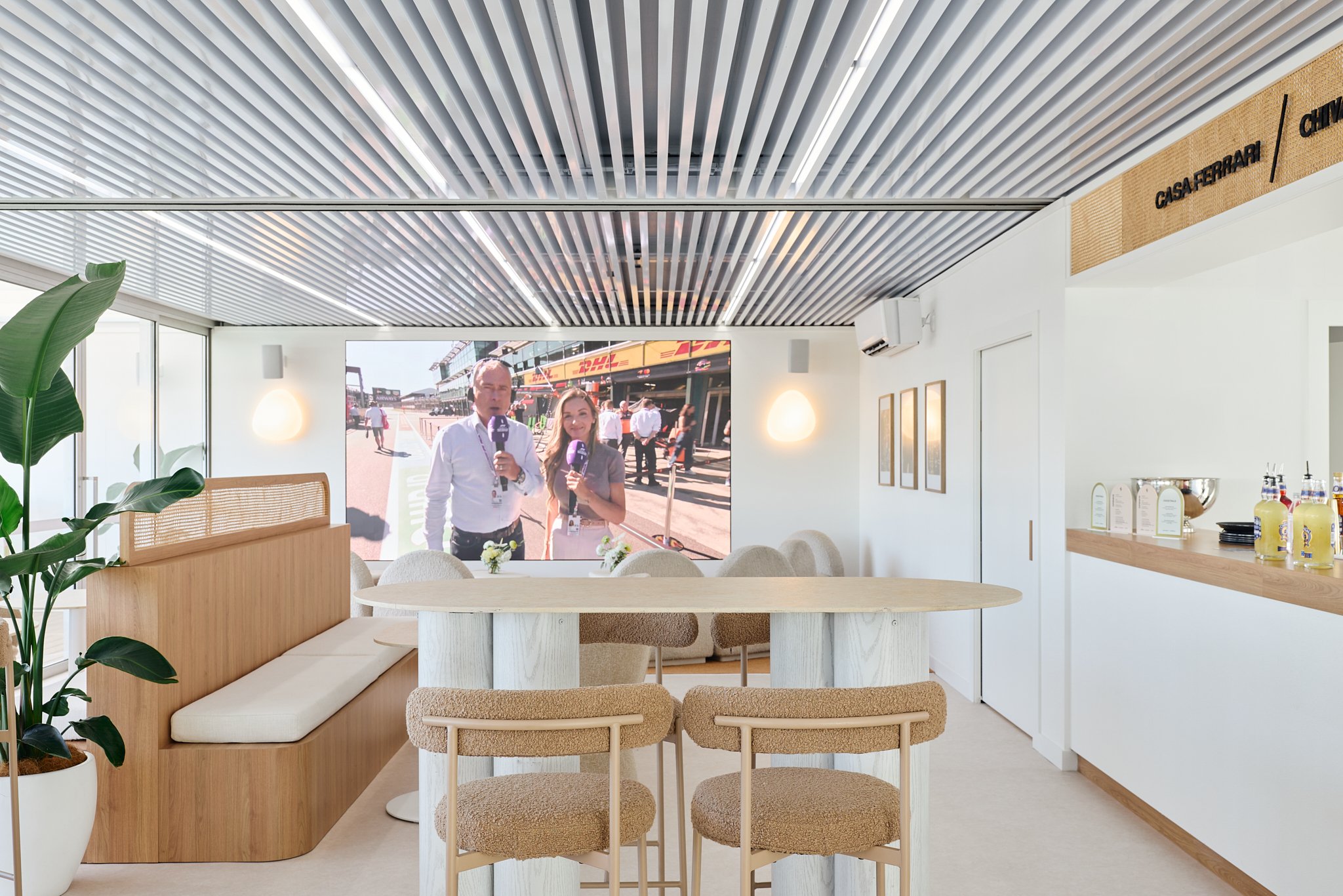 Modern lounge or conference room with seating and a large screen showing two news reporters at a racing event.