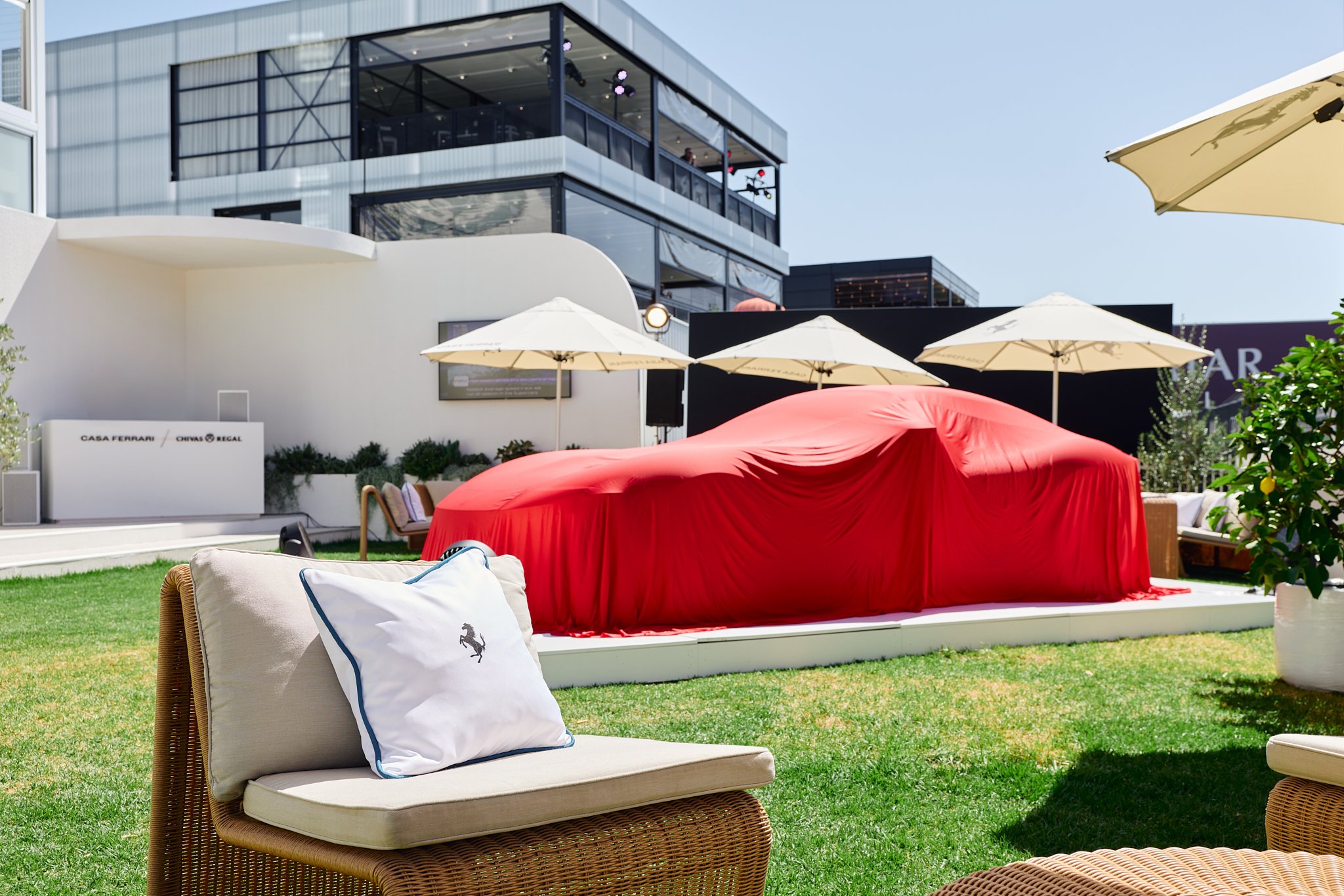 Outdoor patio with wicker furniture, beige cushions, and a white pillow with a black horse logo, surrounded by green grass, white umbrellas, and a covered car draped in red fabric.