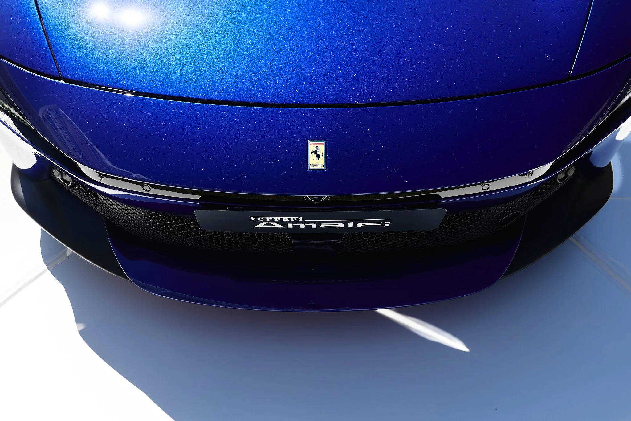 Close-up of a blue Ferrari sports car front, showing the prancing horse emblem on the hood and a Ferrari Anailri badge on the front grille.