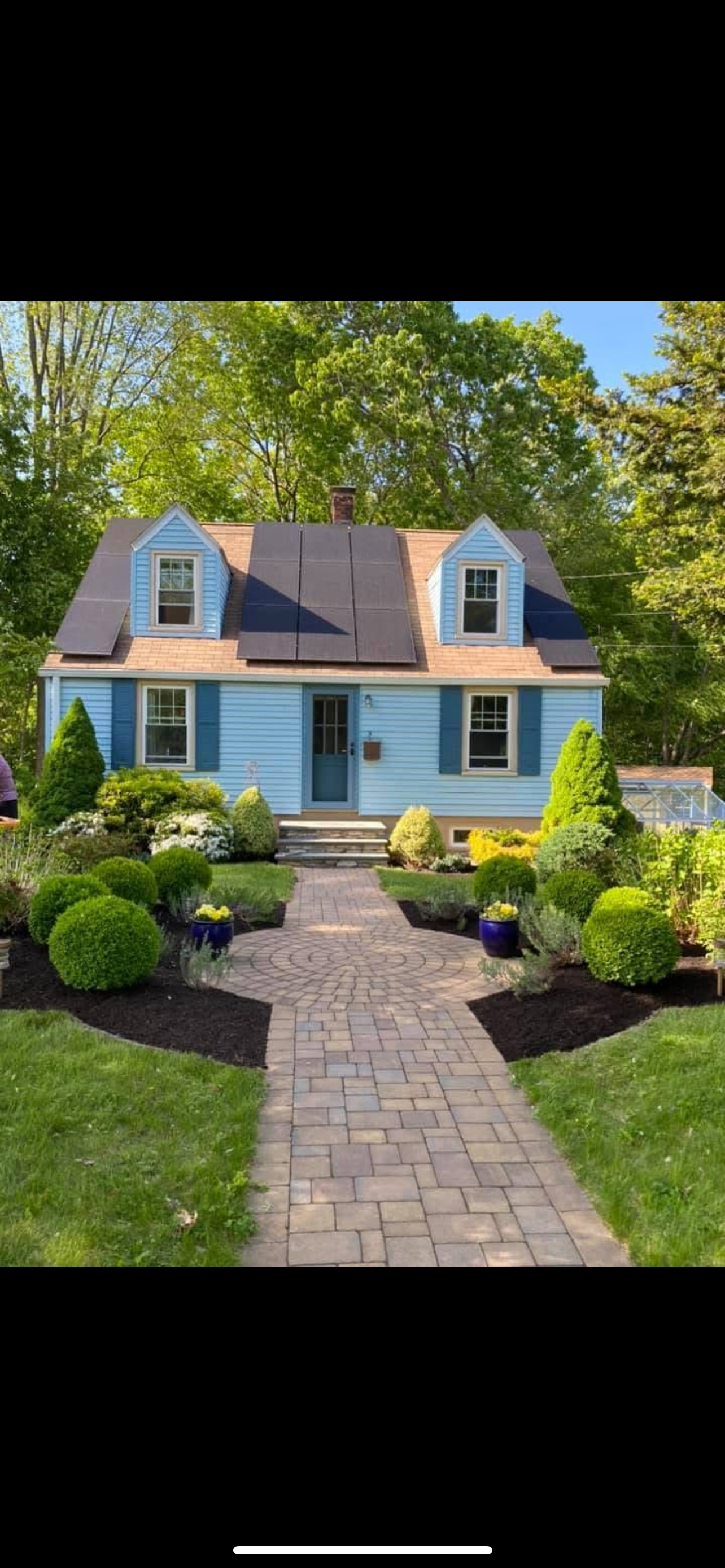 A two-story house painted light blue with dark blue shutters and a smaller attached greenhouse or sunroom on the right. The roof is partially covered with black solar panels. The front yard has a brick walkway leading to the front door, surrounded by