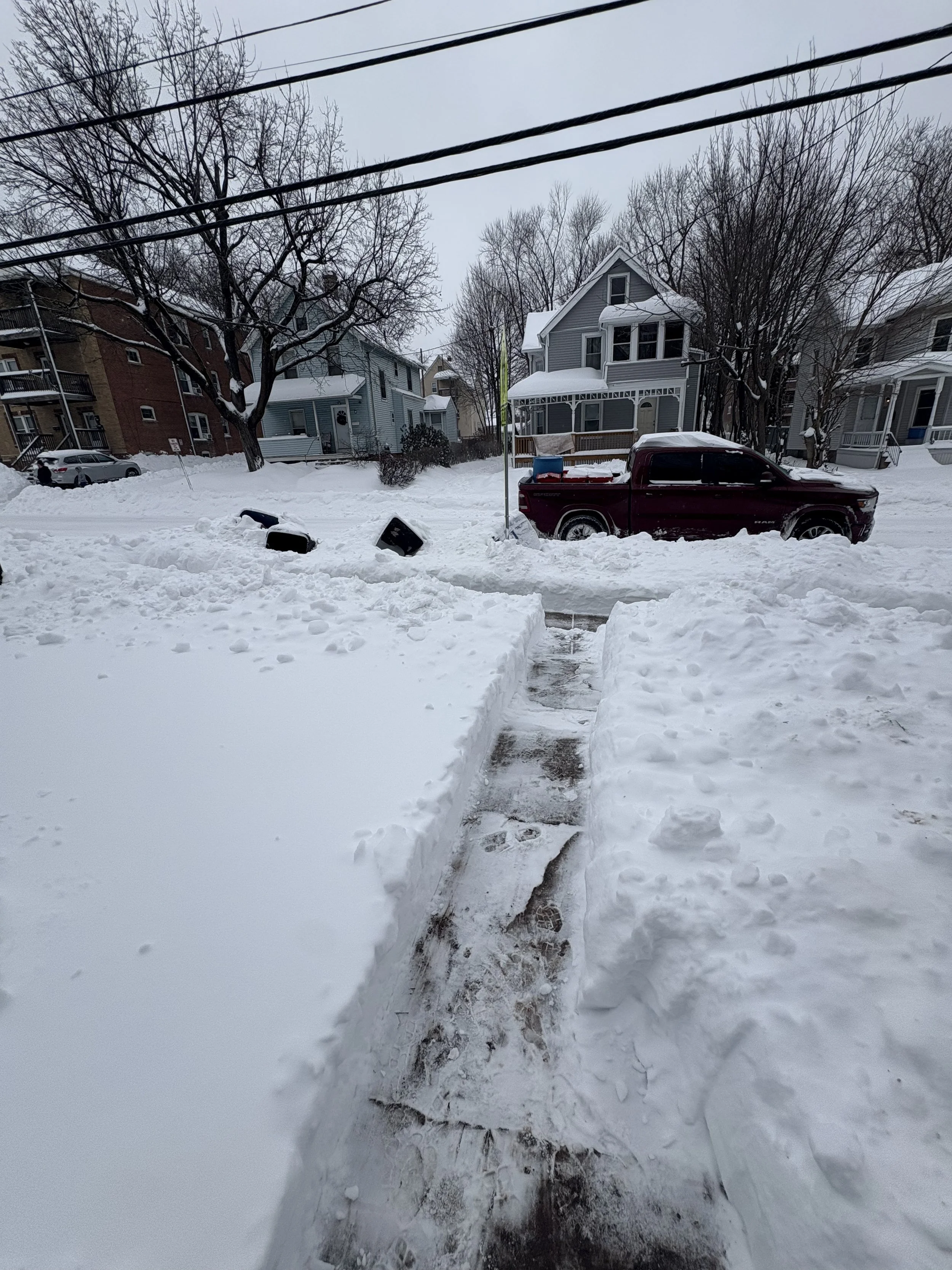 Snow-covered residential street with houses, trees, a red pickup truck, a sidewalk, and a path cleared through the snow.