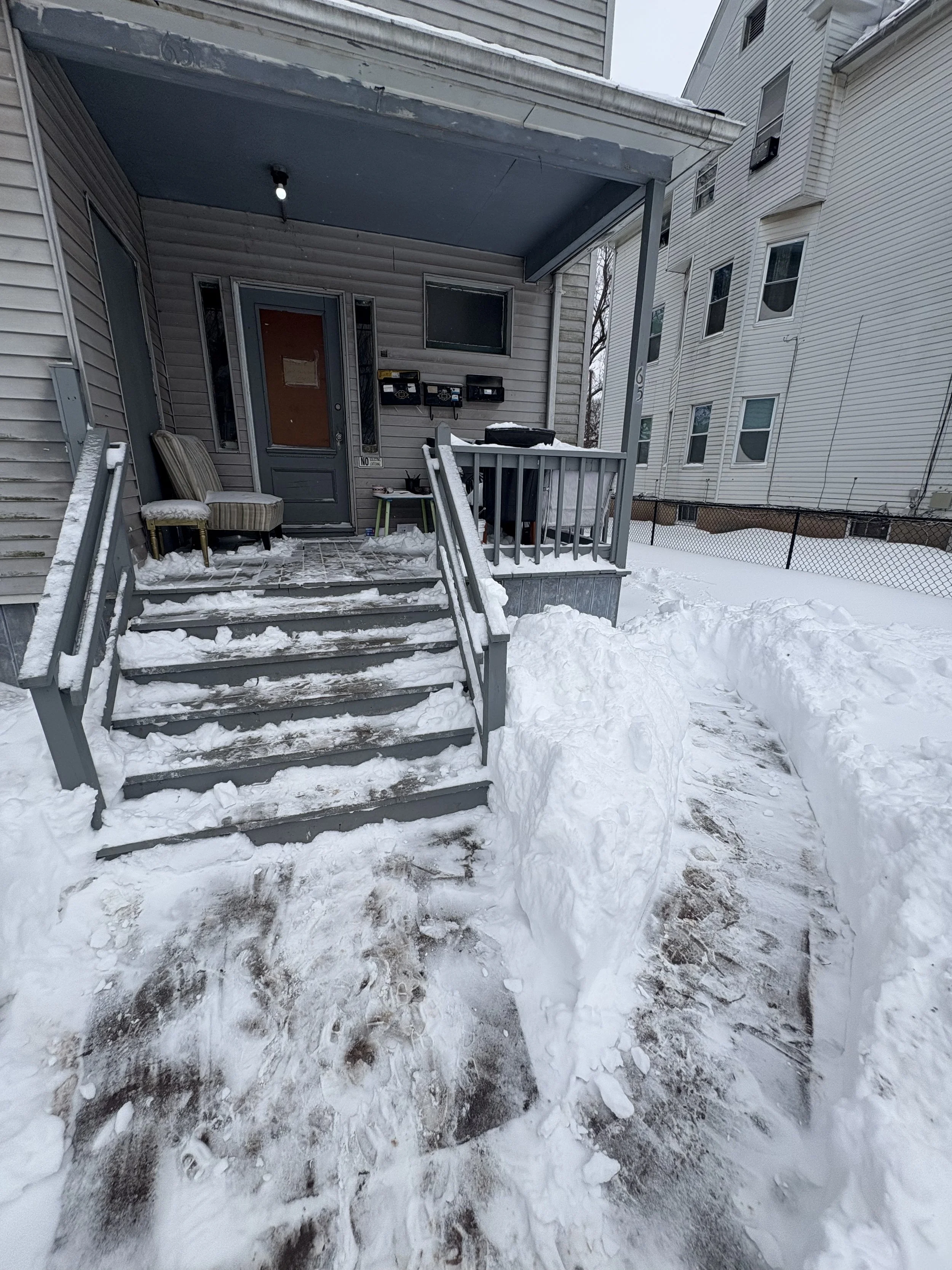A snow-covered front porch with stairs, two outdoor chairs, and a barbecue grill, with snow on the ground and an adjacent building in the background.