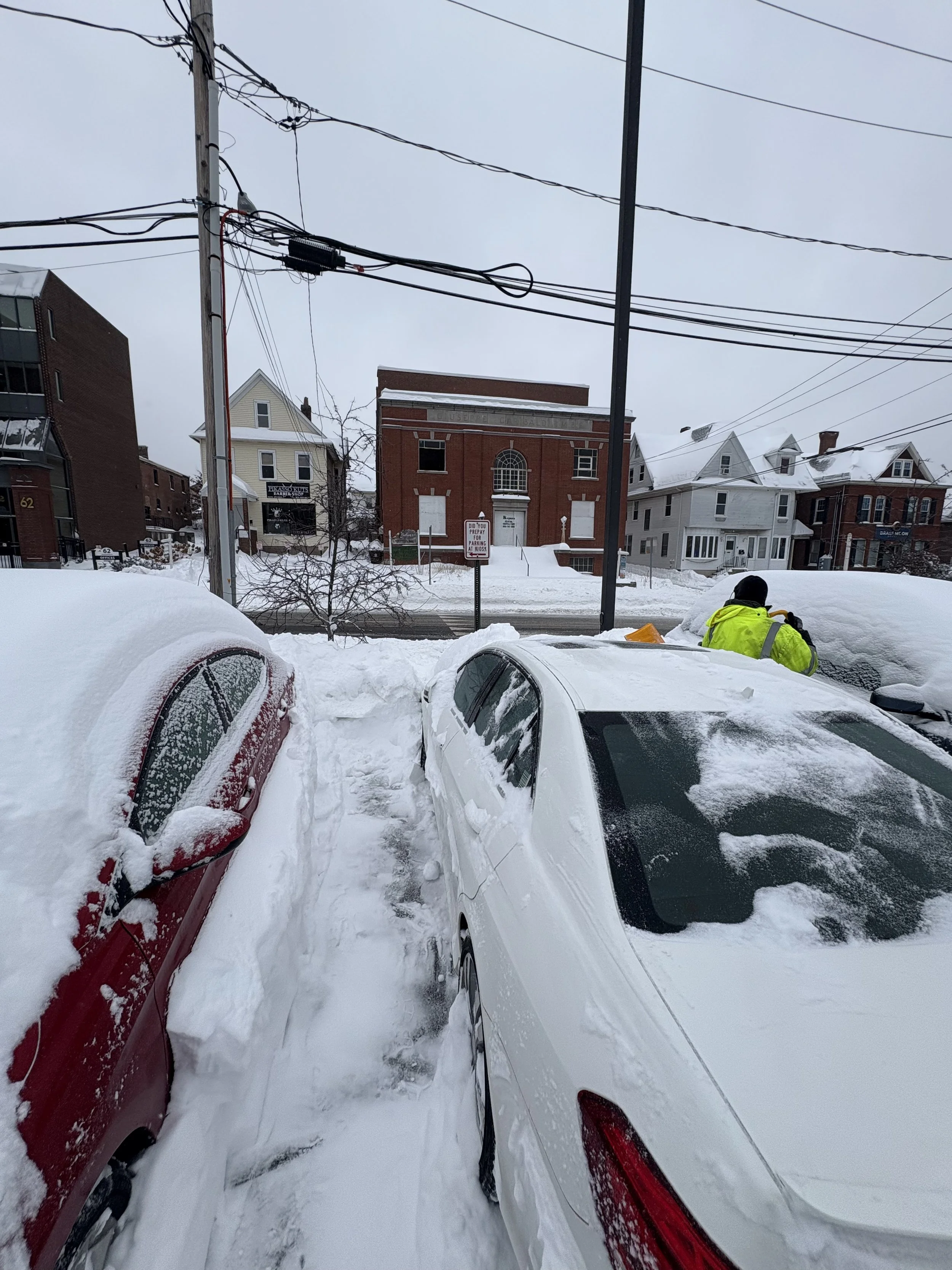 Snow-covered cars parked closely together with a person in a high-visibility jacket shoveling snow nearby, and a background of buildings and power lines in a snowy urban setting.