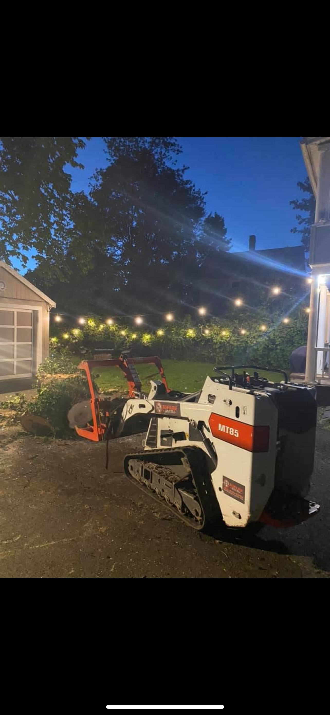 A small tracked excavation machine in a backyard at dusk with string lights overhead, trees in the background, and a house visible on the right.