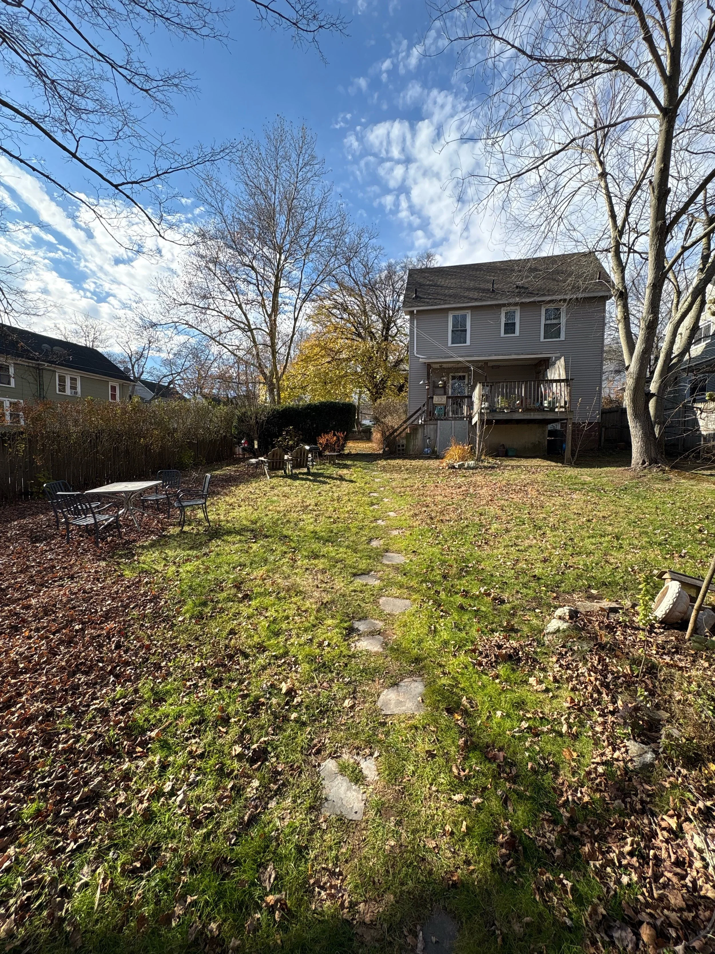 A backyard with a stone pathway leading to a house with a small wooden deck. There are four black outdoor chairs around a table on the left, bare trees, and a mix of green grass and fallen leaves on the ground. The sky is mostly clear with some cloud