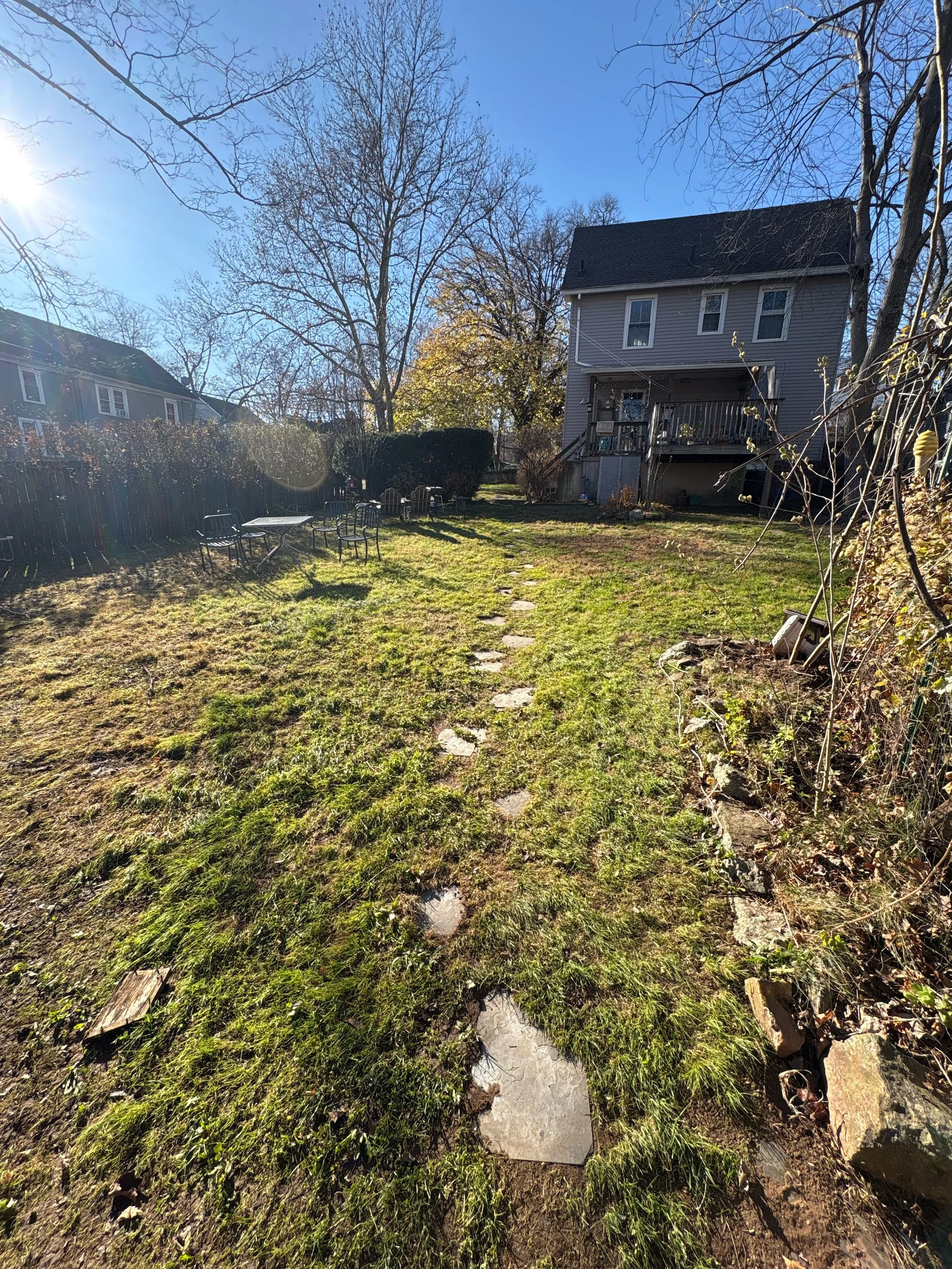 A backyard with a stone pathway and outdoor furniture, surrounded by grass and trees with no leaves, under a clear blue sky with the sun shining.