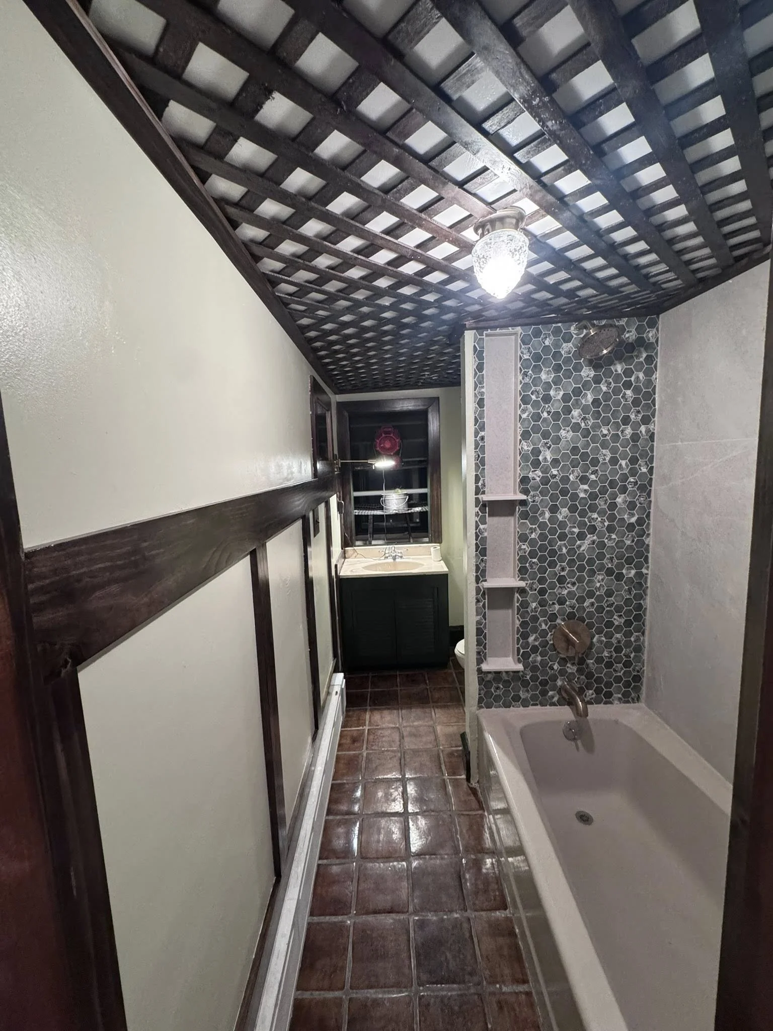 A small bathroom with a bathtub, a shower with dark hexagon tiles, a small sink, and a window with shelves.