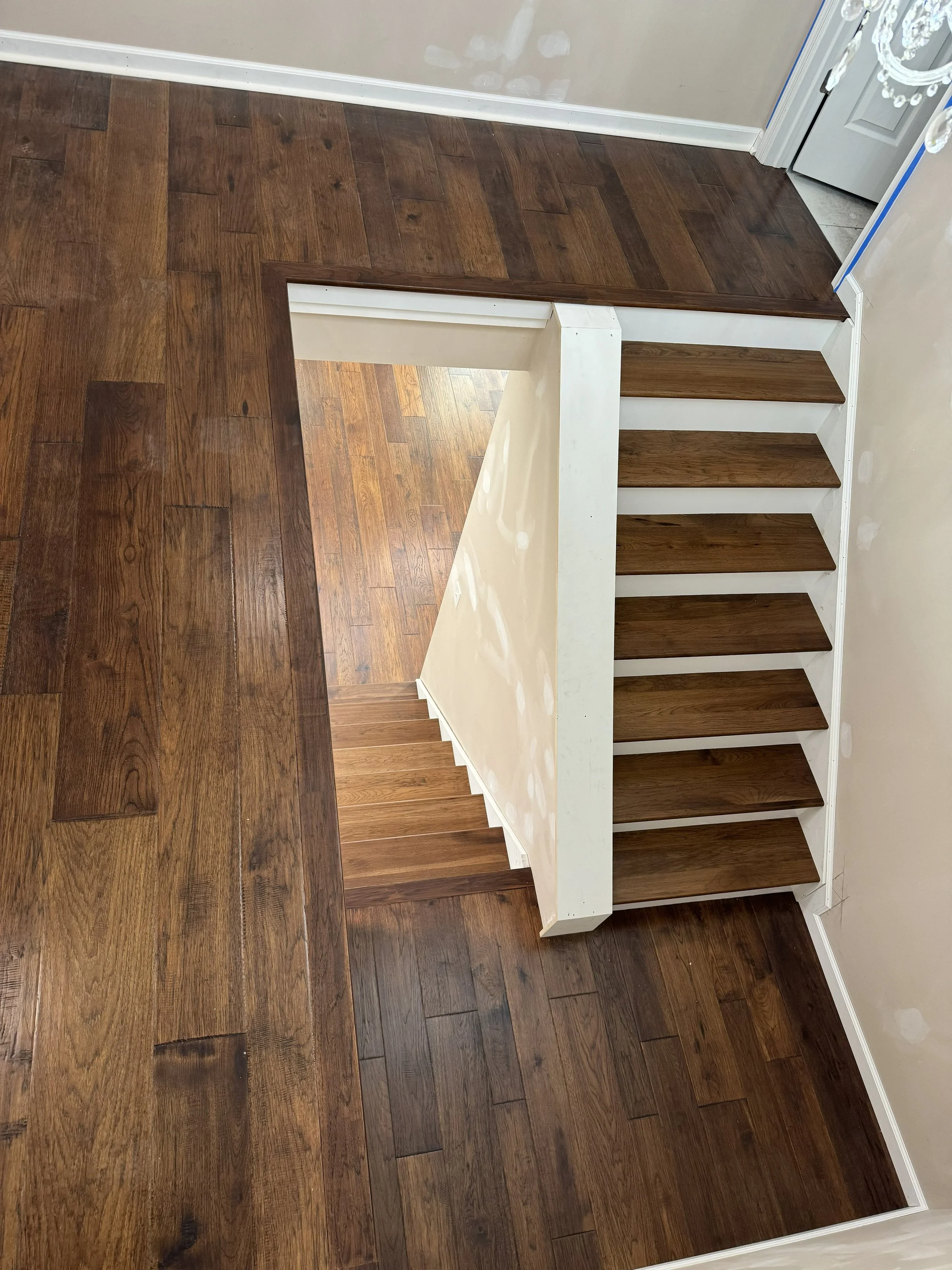 Empty staircase with wooden steps, white risers, and a white wall with paint patches, leading to a lower level with hardwood flooring. Part of a chandelier is visible at the top right corner.