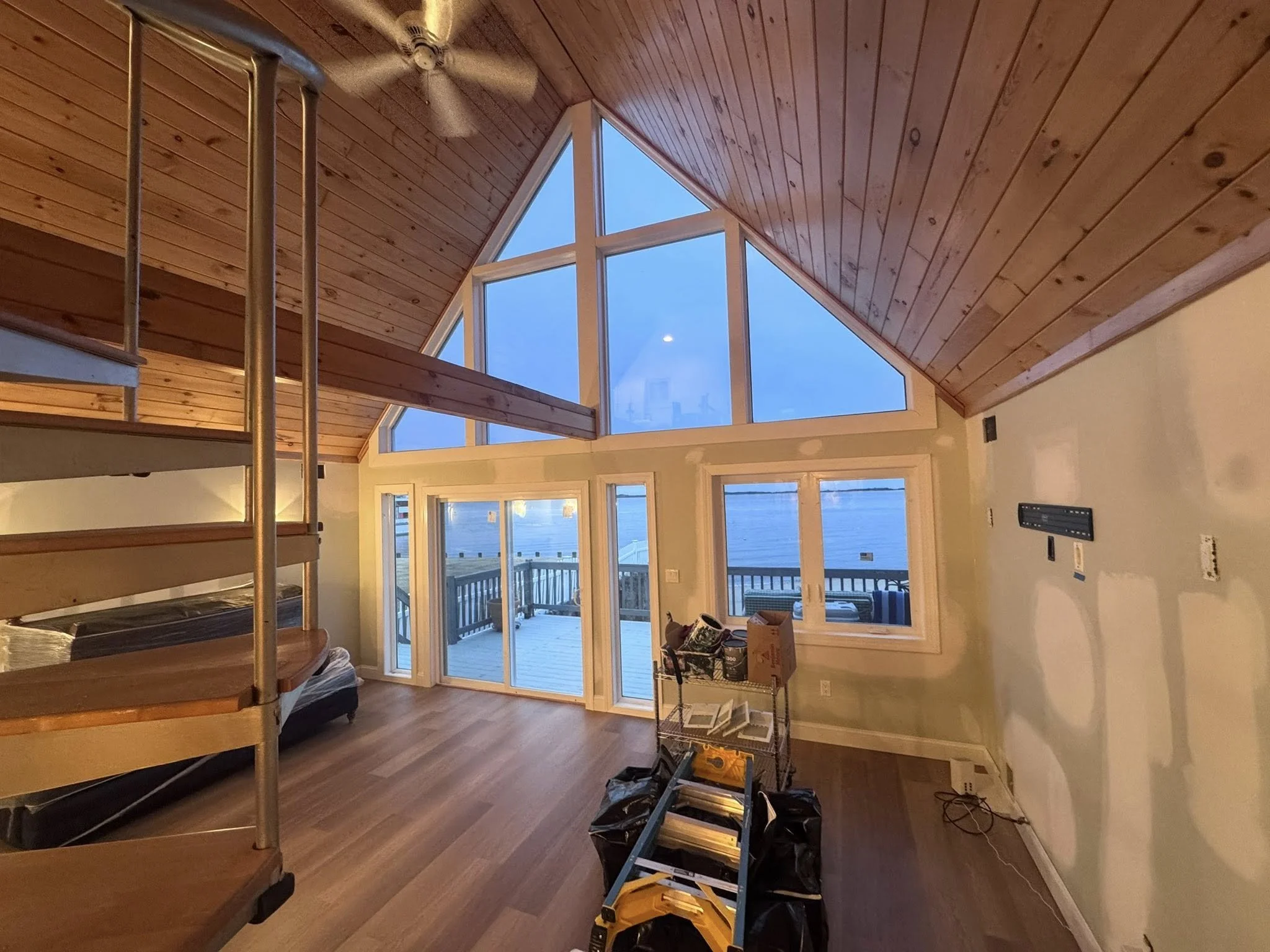 Interior of a house with wooden ceiling, large windows with ocean view, and unfinished walls, with tools and construction materials on the floor.
