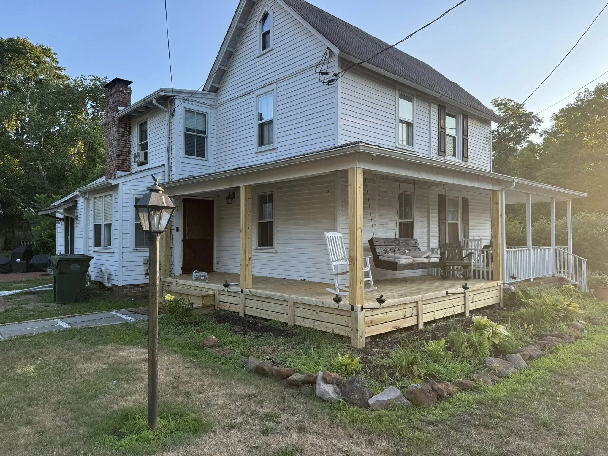 A two-story white house with a wooden porch under construction, outdoor furniture, a brick chimney, and a landscaped garden with rocks and plants, surrounded by trees.