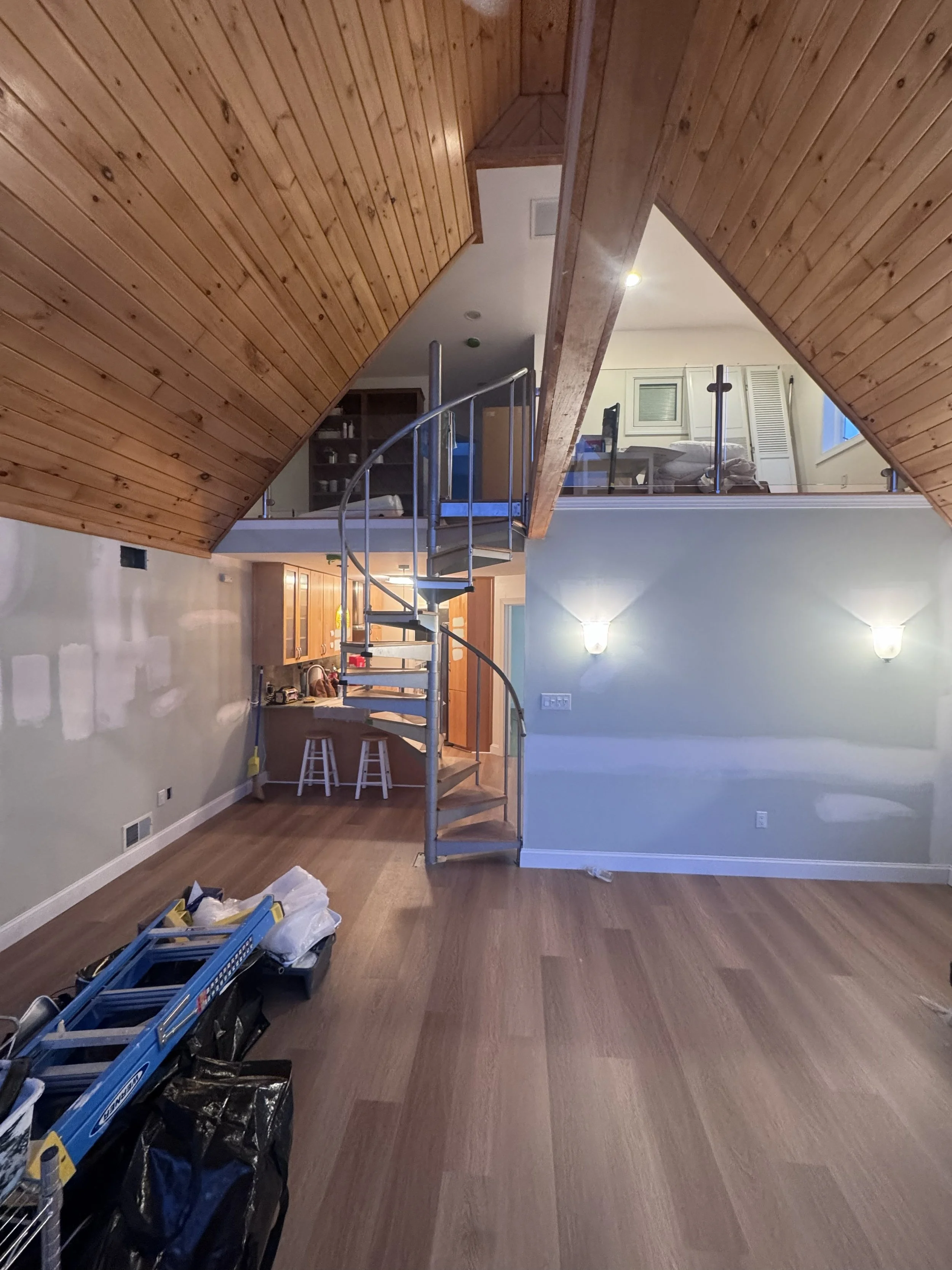 Interior of a house with a spiral staircase leading to a loft area, wood-paneled ceiling, and a living space with some tools and supplies on the floor, walls with light fixtures, and a kitchen area with bar stools.