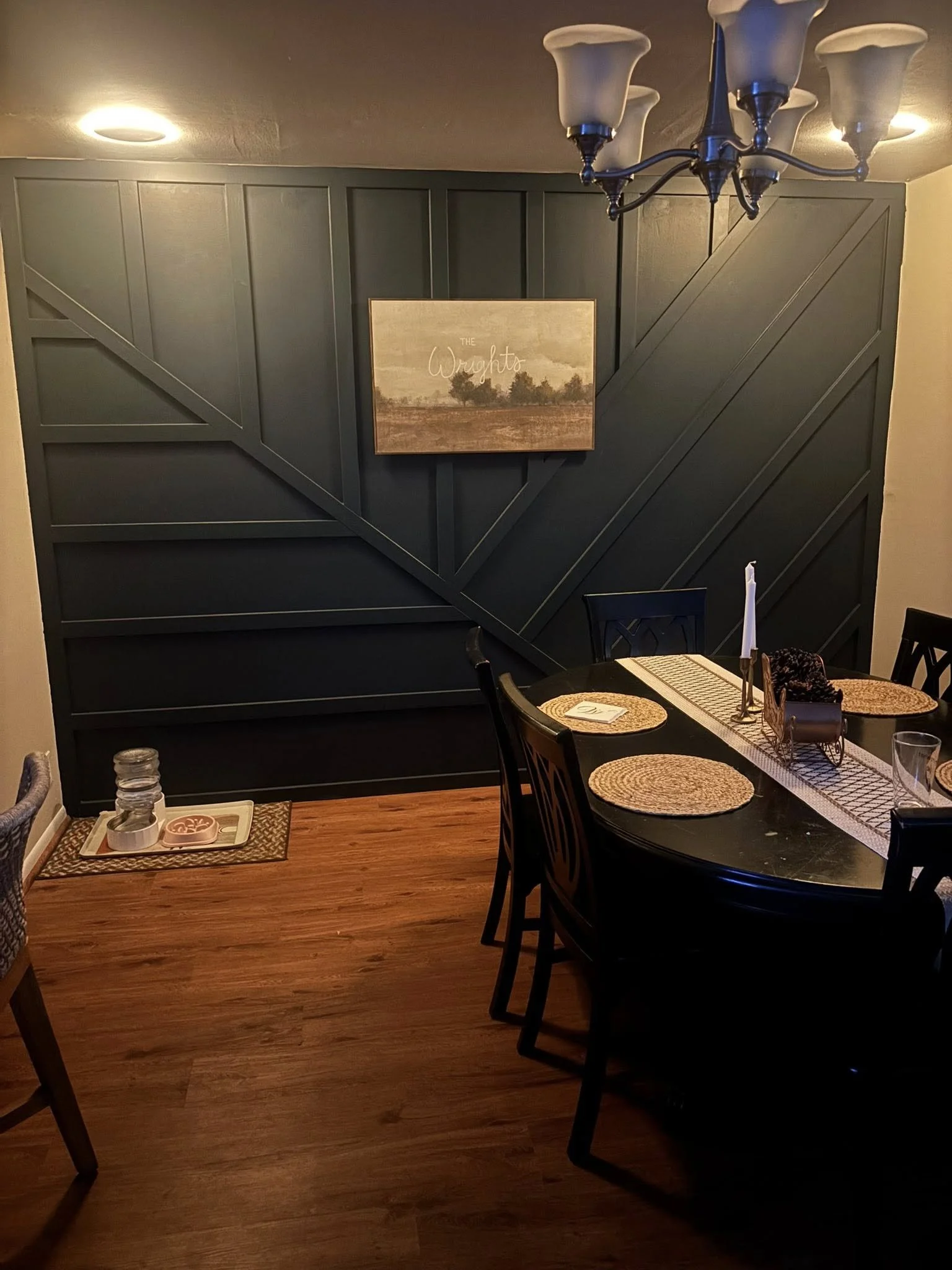 Dining room with black table and chairs, dark accent wall with wooden trim, table set with placemats, candlestick, and decor, chandelier overhead, hardwood floor.