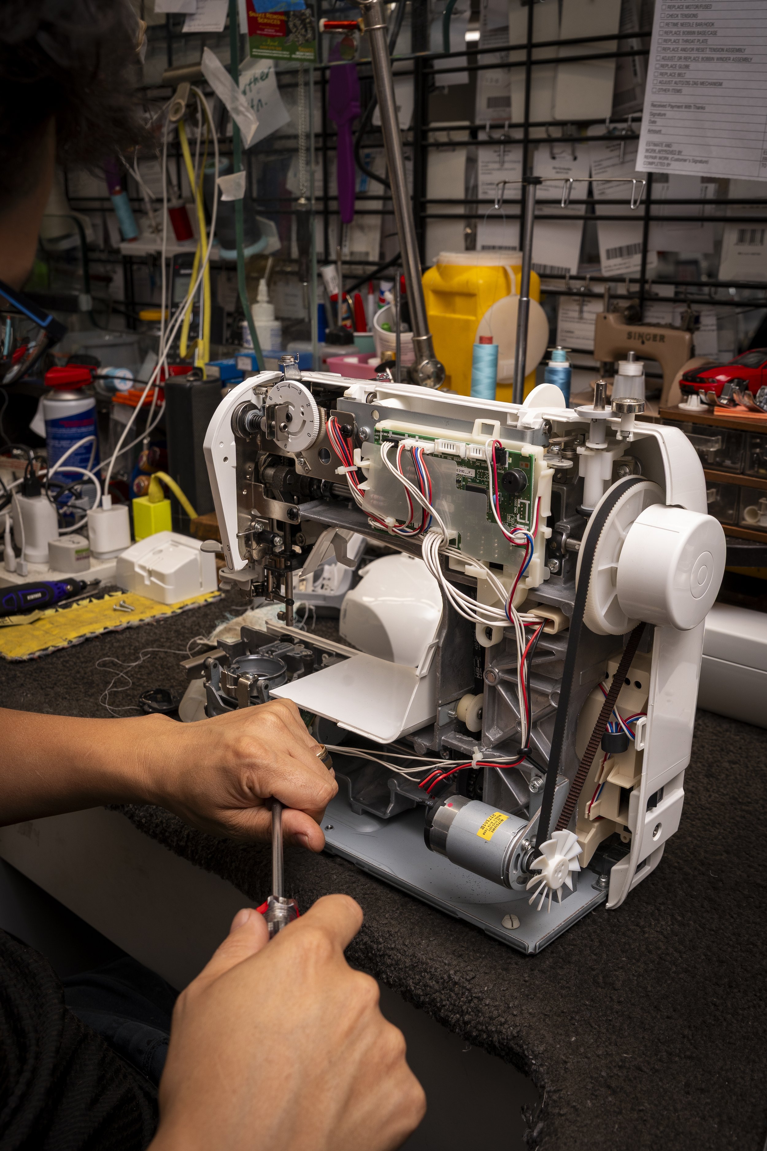 Person repairing a sewing machine on a workbench surrounded by tools and supplies in a workshop.