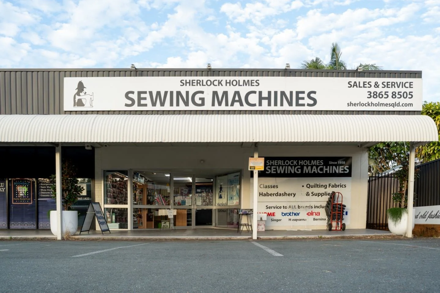 Storefront sign for Sherlock Holmes Sewing Machines with contact information, and an entrance displaying sewing supplies and machines.