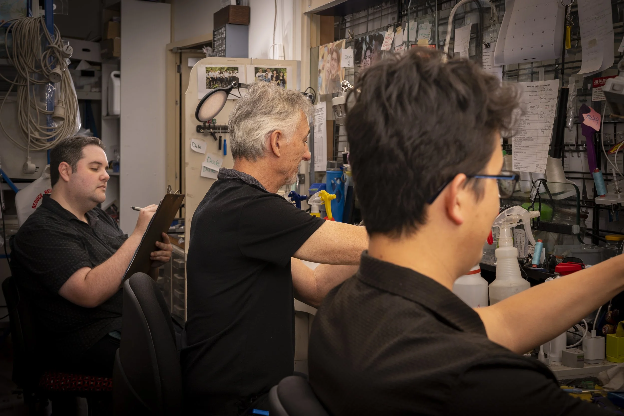 Three men working on sewing machines in a workshop, surrounded by tools, notes, and photos on the wall.