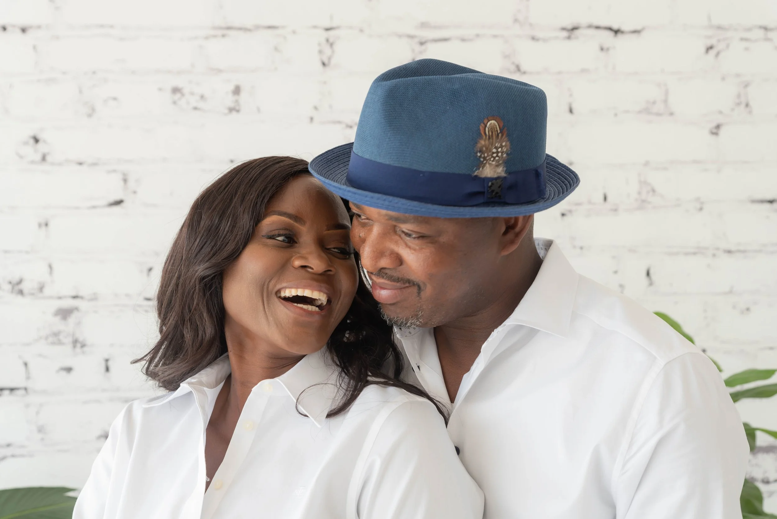 A happy Black woman and an older Black man in white shirts sharing an intimate moment, with their foreheads touching and smiling, against a white brick wall background.