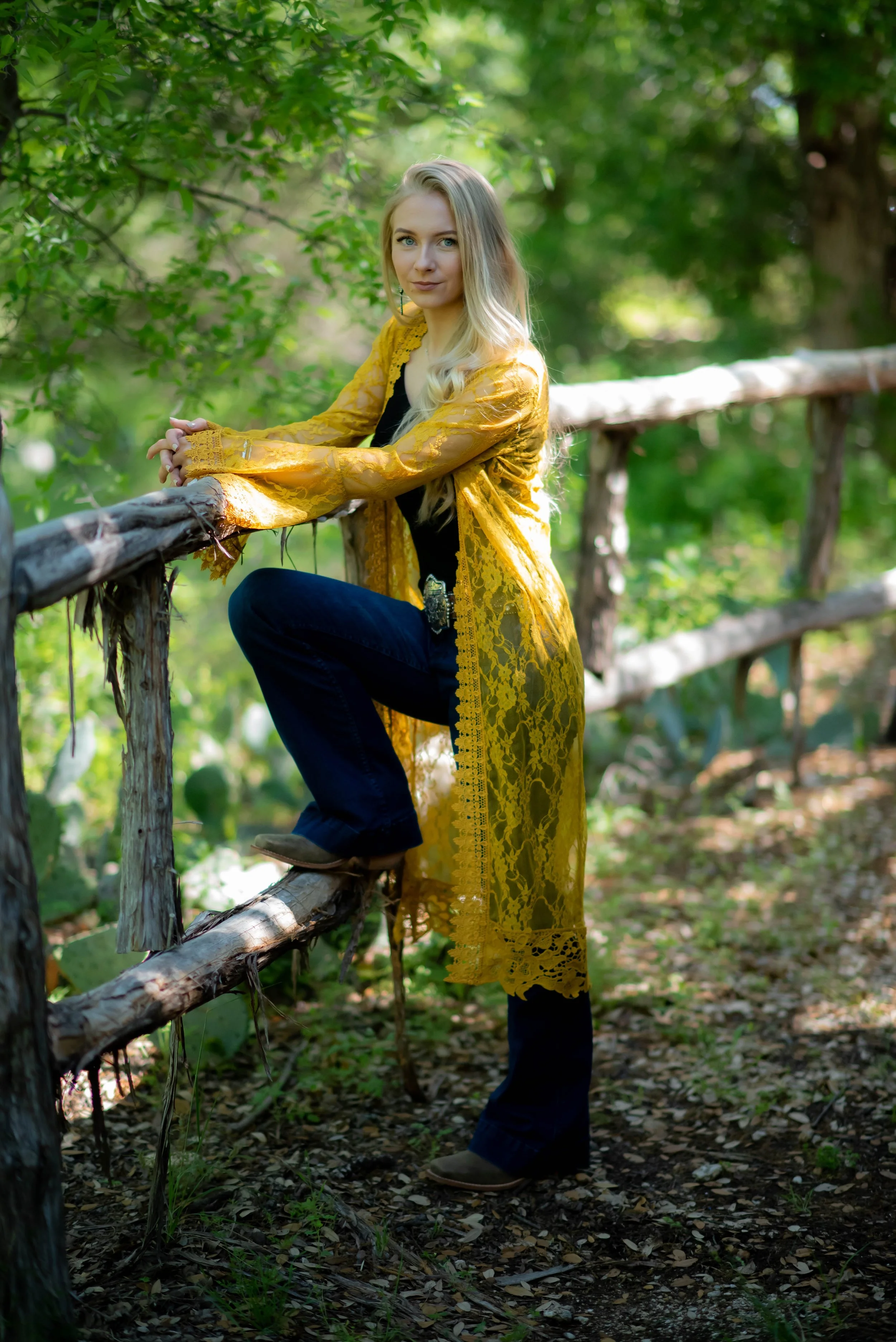 A young woman with long blonde hair, wearing a yellow lace kimono over black clothing, is standing outdoors on a rustic wooden bridge surrounded by lush green trees.
