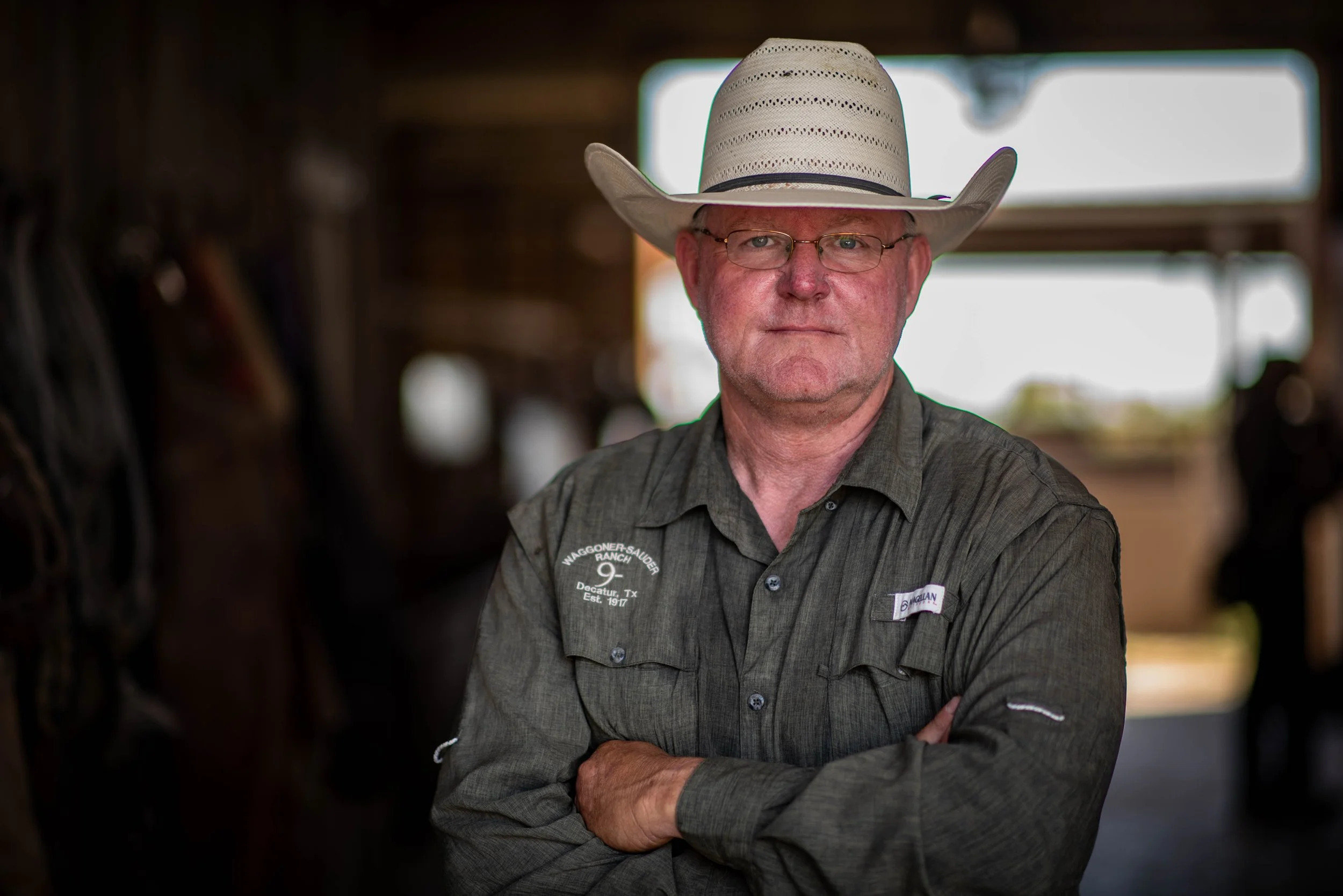 A man wearing glasses and a cowboy hat standing with crossed arms, wearing a gray button-up shirt with a logo and text on the chest pocket, in a rustic setting with blurred background.