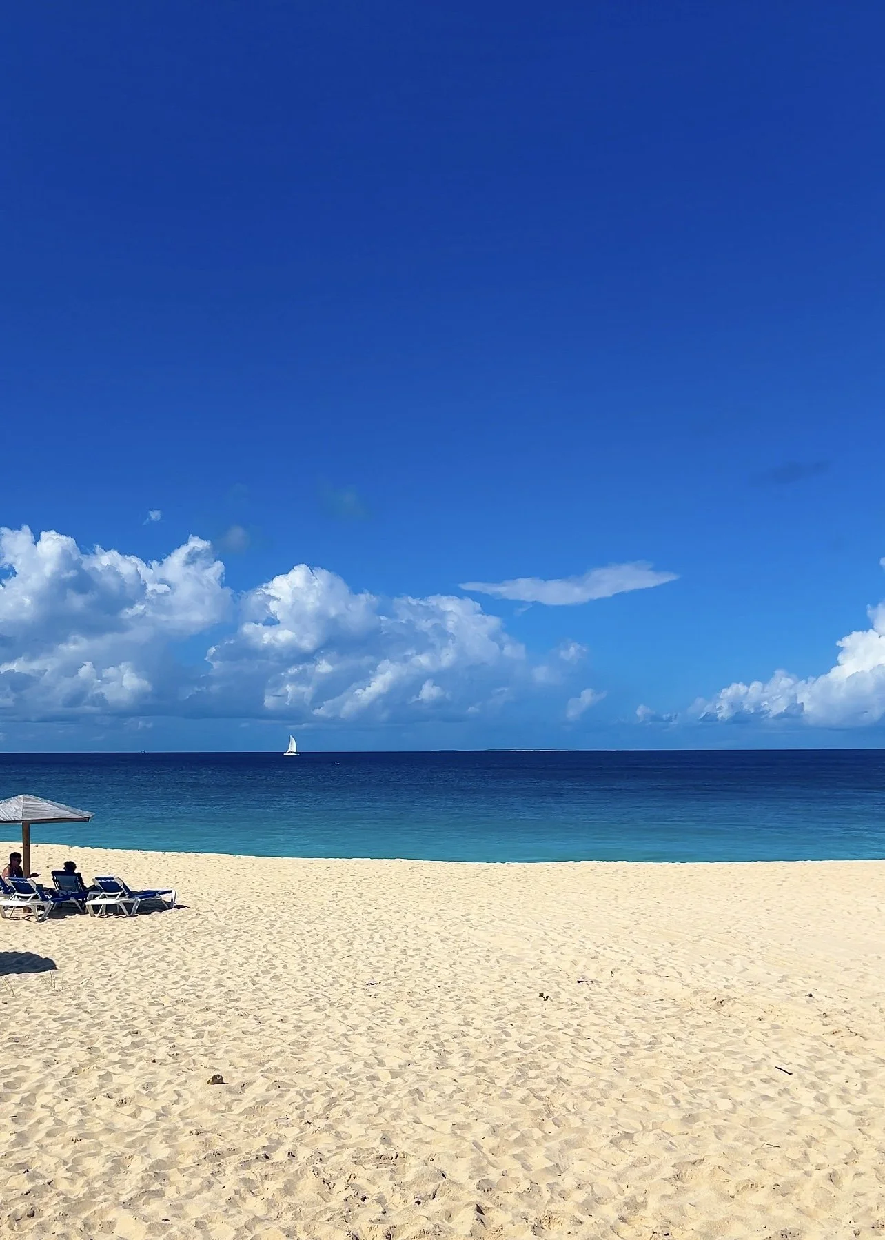 A sandy beach with beach chairs and a large umbrella on the left, blue ocean water, a sailboat in the distance, and a partly cloudy sky with white clouds.