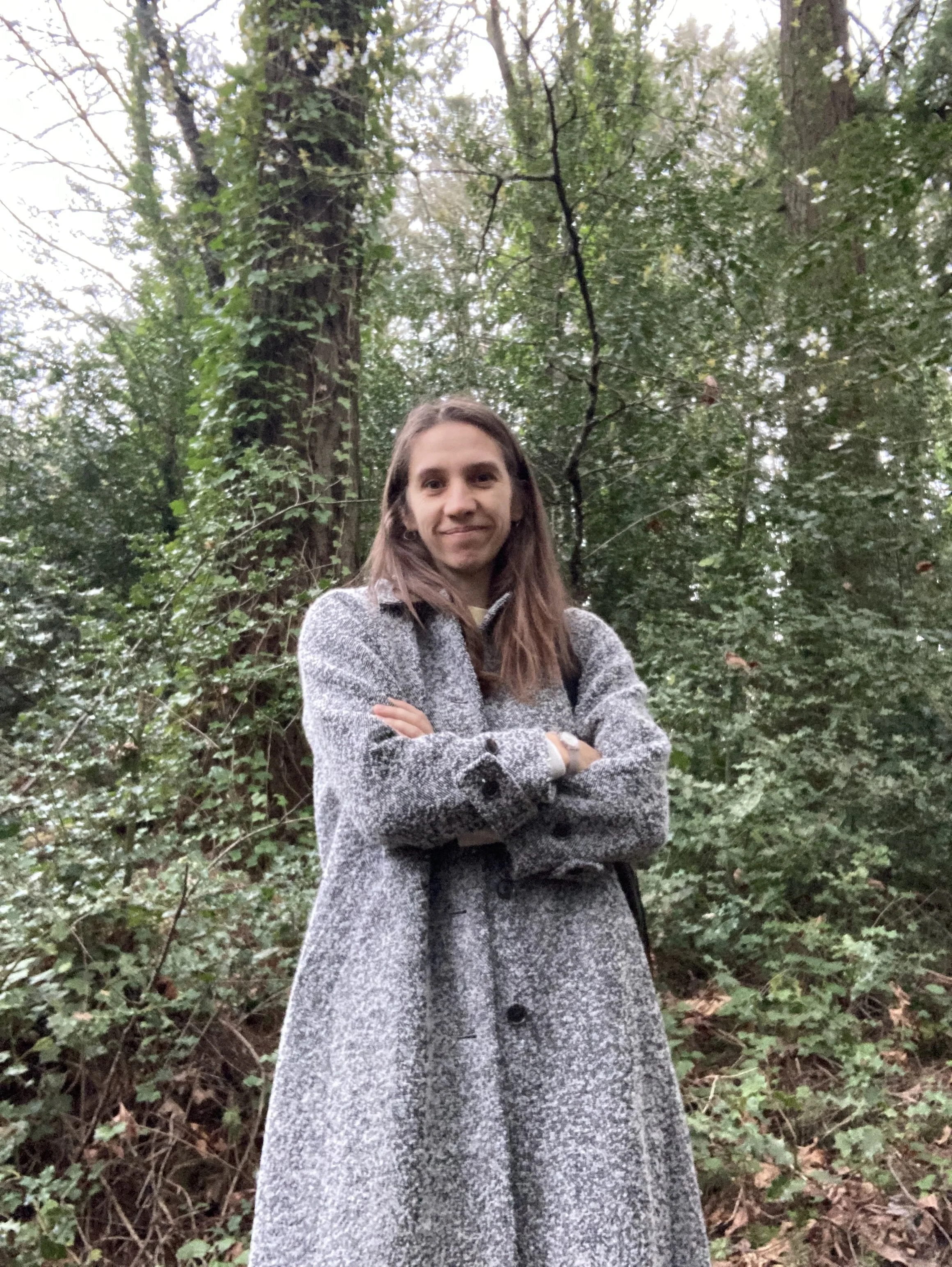 A woman in a gray coat standing with arms crossed in a wooded area with tall trees and green foliage.
