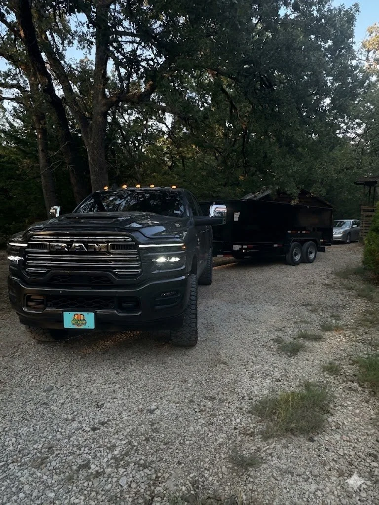 A black RAM pickup truck towing a large black trailer, parked on a gravel driveway surrounded by trees and a few other vehicles in the background.