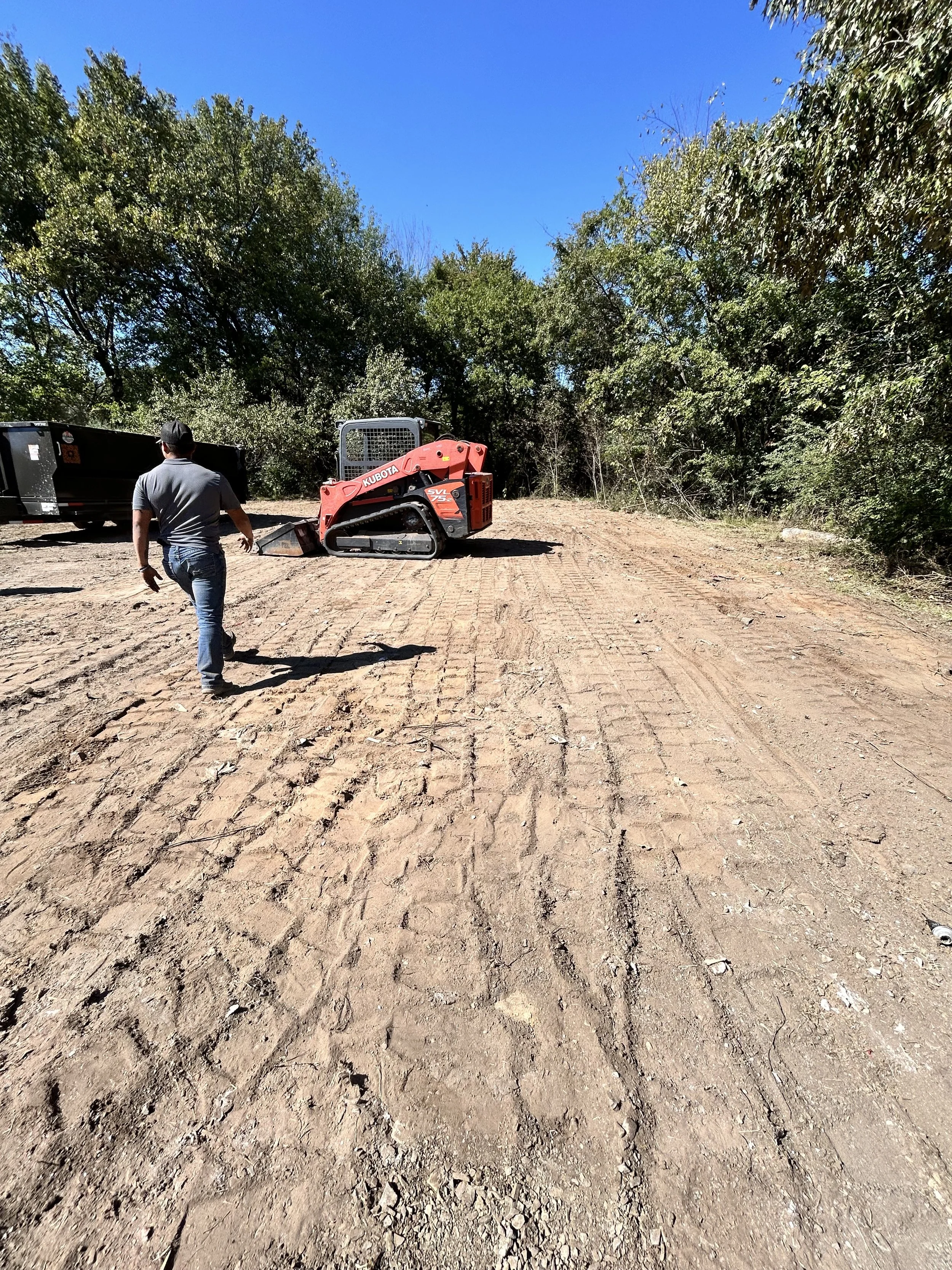 A man walking on a dirt construction site with a small red Kubota excavator and a trailer in the background, trees lining the area under a clear blue sky.
