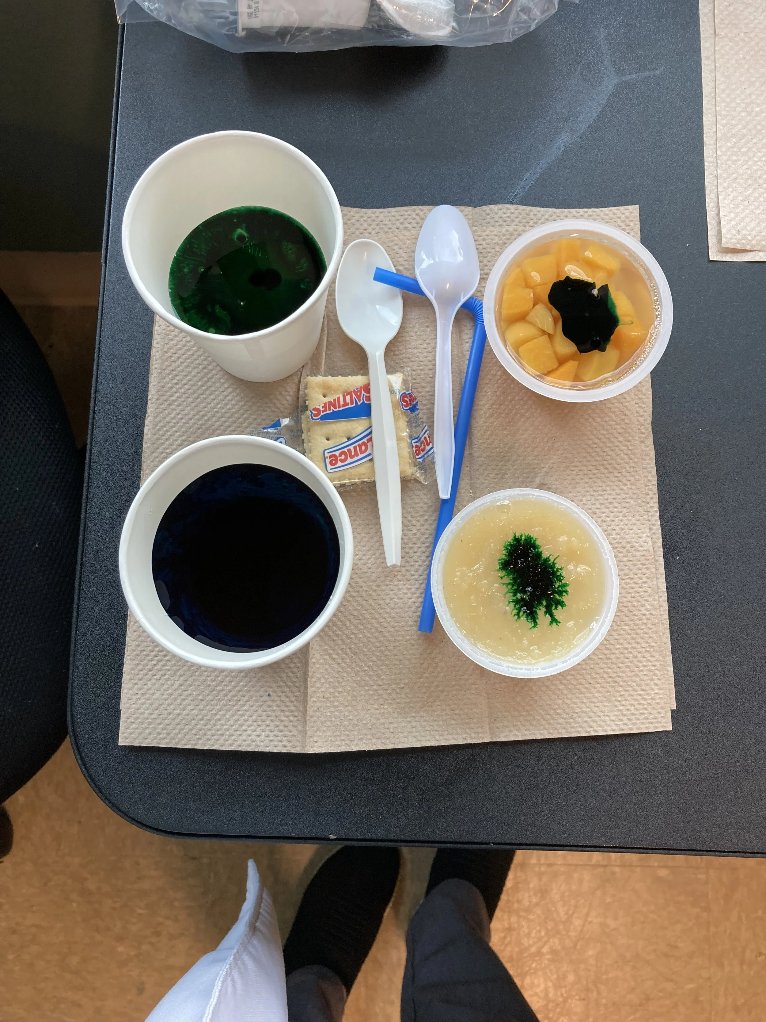 A table with a light brown paper napkin, holding a container of chopped yellow peaches in syrup, a container of yellow pudding with green and black garnish, and a small container of dark-colored beverage. There are two plastic spoons, one white and one blue, and a white and blue packaged cracker.