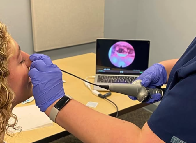 Medical professional performs an oral exam on a patient using a small camera, with a laptop displaying an internal view on the screen.