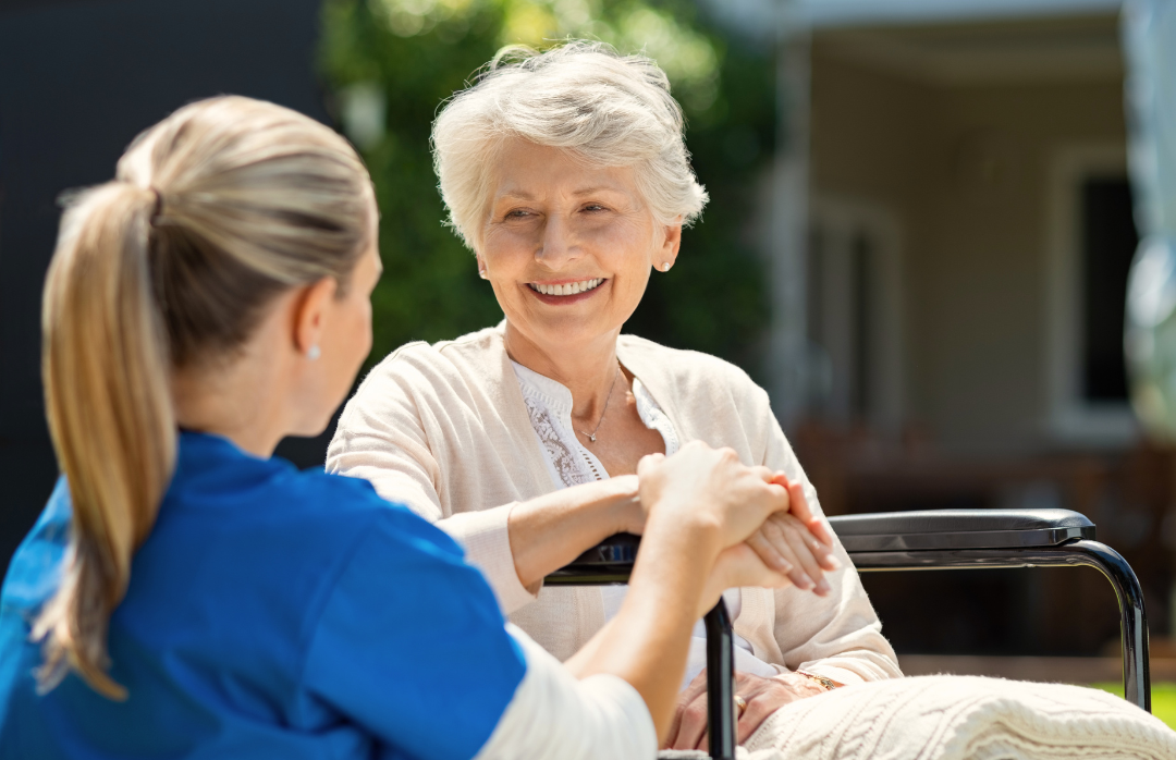 An elderly woman smiling and holding hands with a caregiver outdoors on a sunny day.