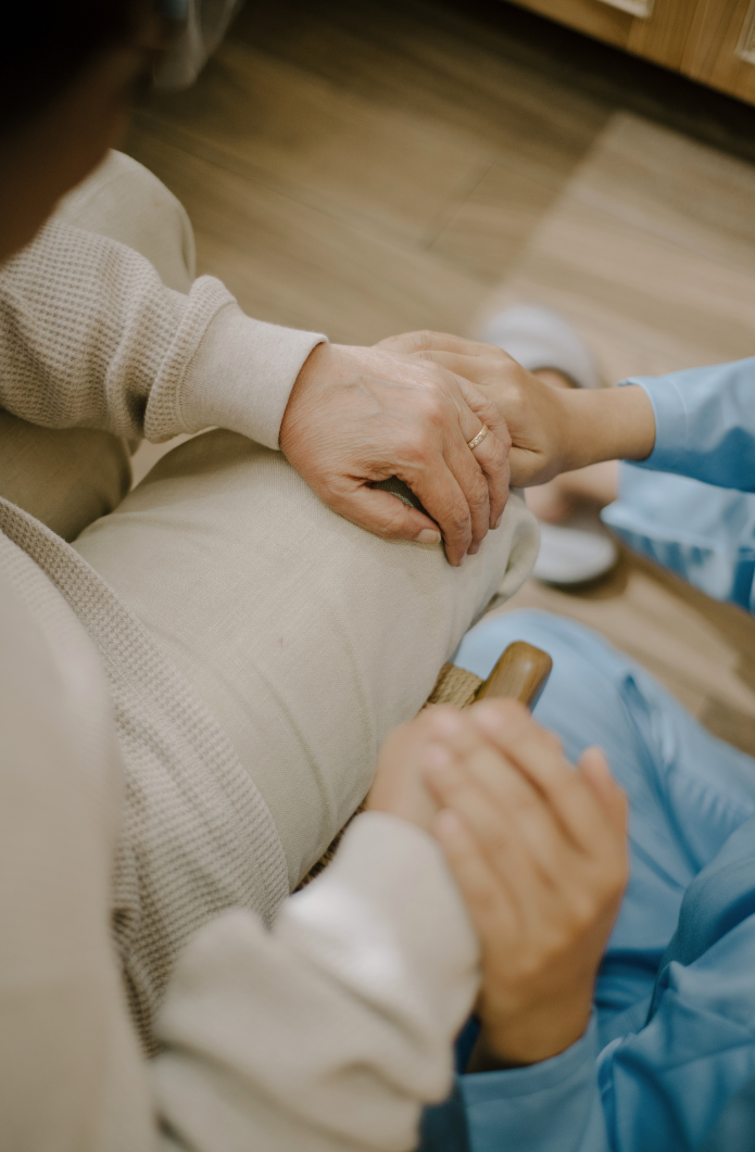 Close-up of two people holding hands, one elderly and one younger, inside a room with wooden flooring.