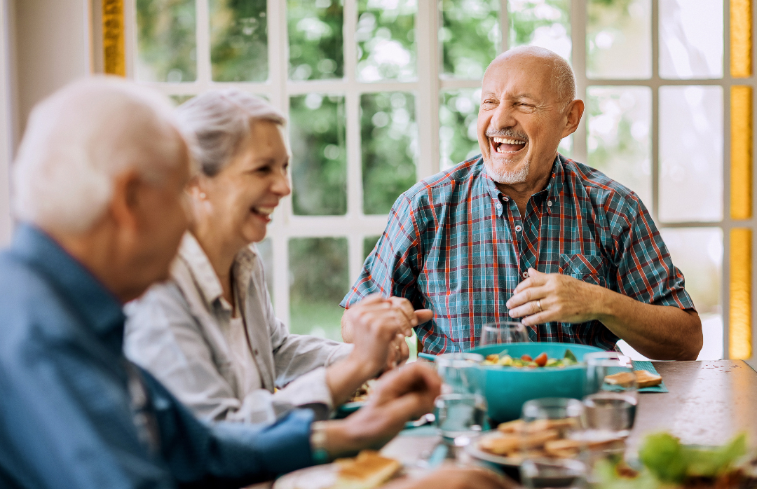 Three older adults around a table, laughing and enjoying a meal together in a bright, sunny room with large window views of greenery.