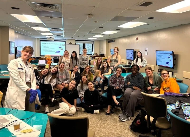 Group of students and instructor in a classroom with multiple computer monitors and a presentation slide about intervention assessment.