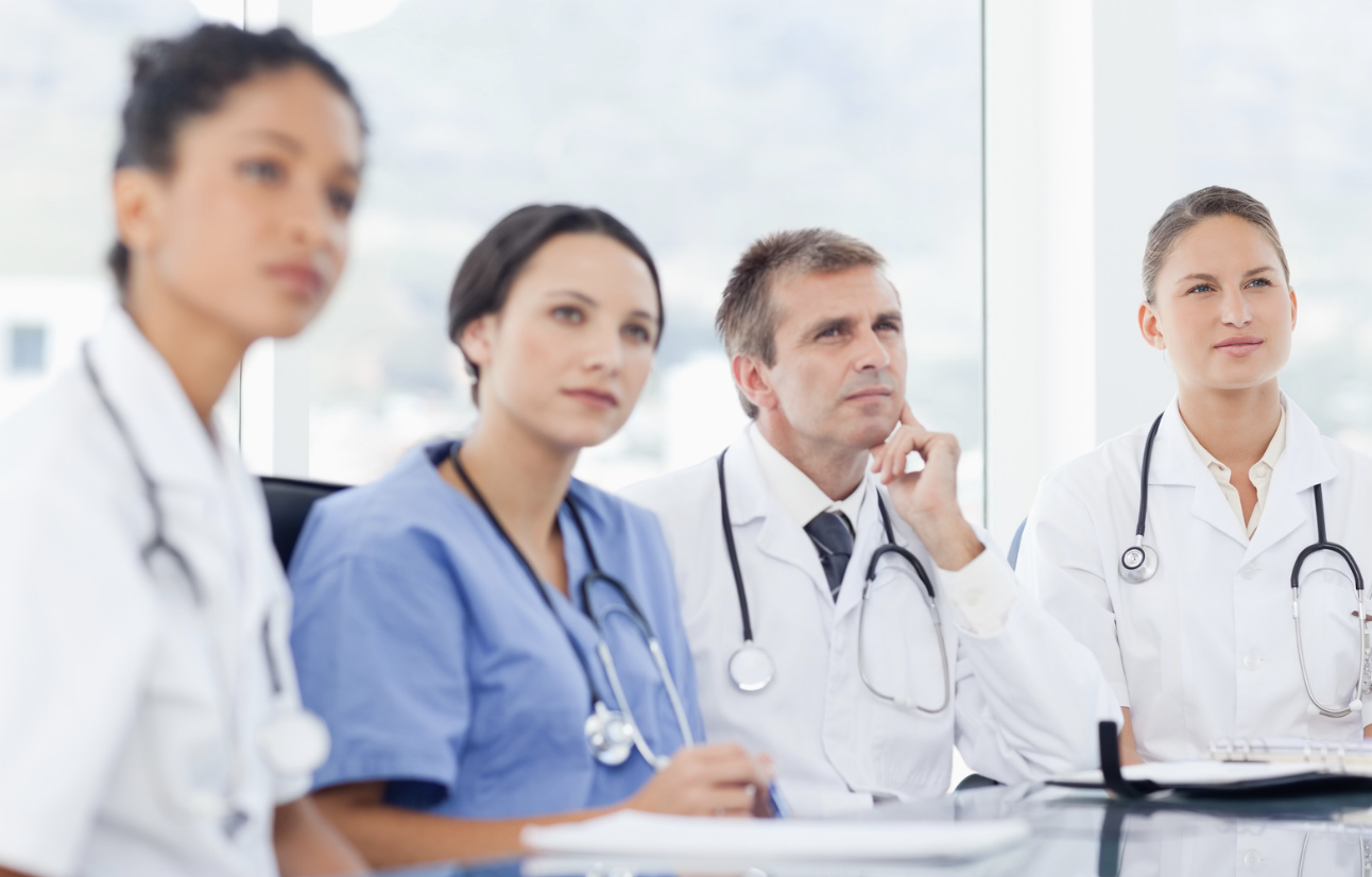 Group of four doctors and healthcare professionals sitting during a meeting in a bright room with large windows, listening attentively.