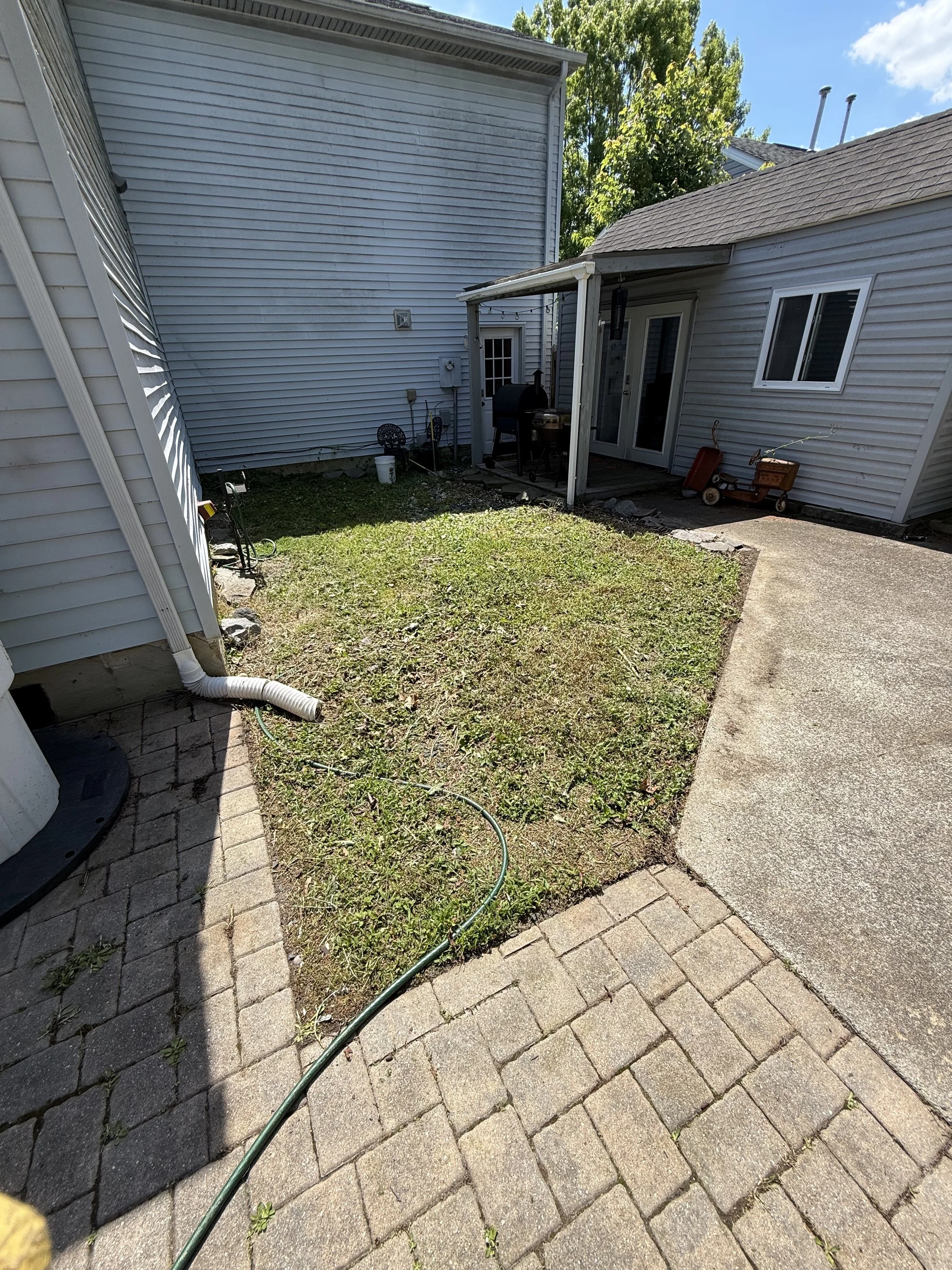 Backyard with small patch of grass, paved patio, white siding house, sliding glass door, outdoor grill, child's wagon, garden tools, and trees.