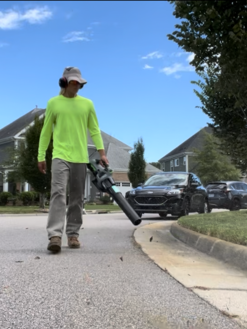 Person wearing a bright yellow-green long sleeve shirt, gray pants, and a hat, using a leaf blower on a suburban sidewalk.