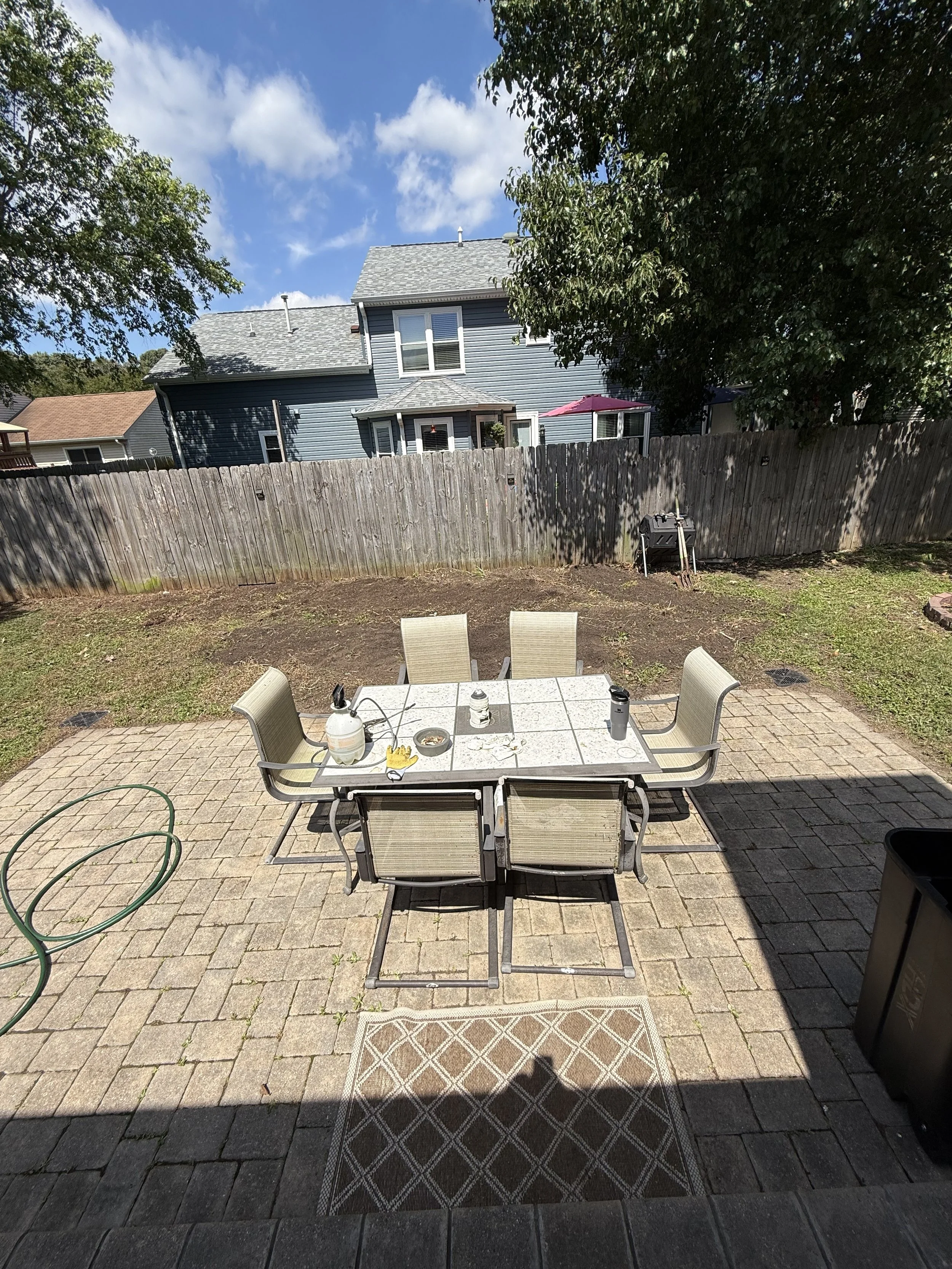 Backyard patio with a table and six chairs, some items on the table including a water bottle, a thermos, and a bowl. There is a garden hose on the ground and a barbecue grill in the background, with a wooden fence and a house with blue siding and a p
