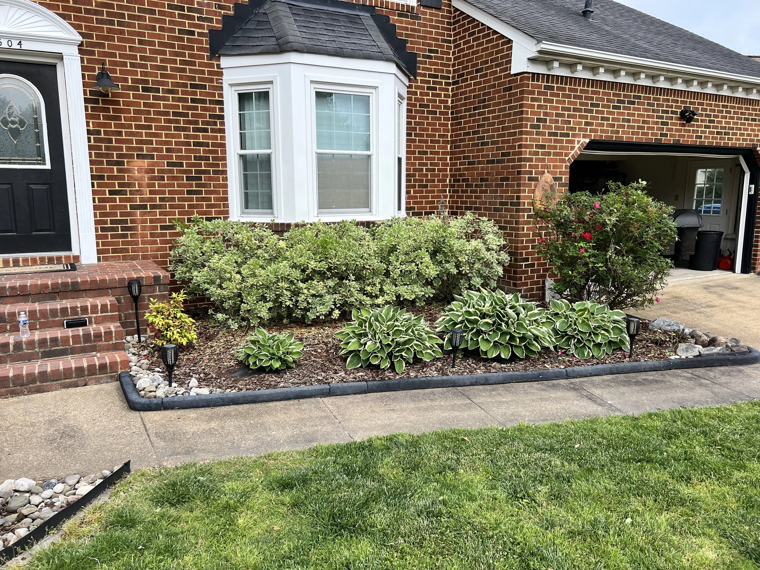 A brick house with a front yard garden containing green bushes, hosta plants, and small decorative lights. The yard is bordered by a black edging and has a sidewalk in front. There are brick steps leading to a black front door on the left and an open