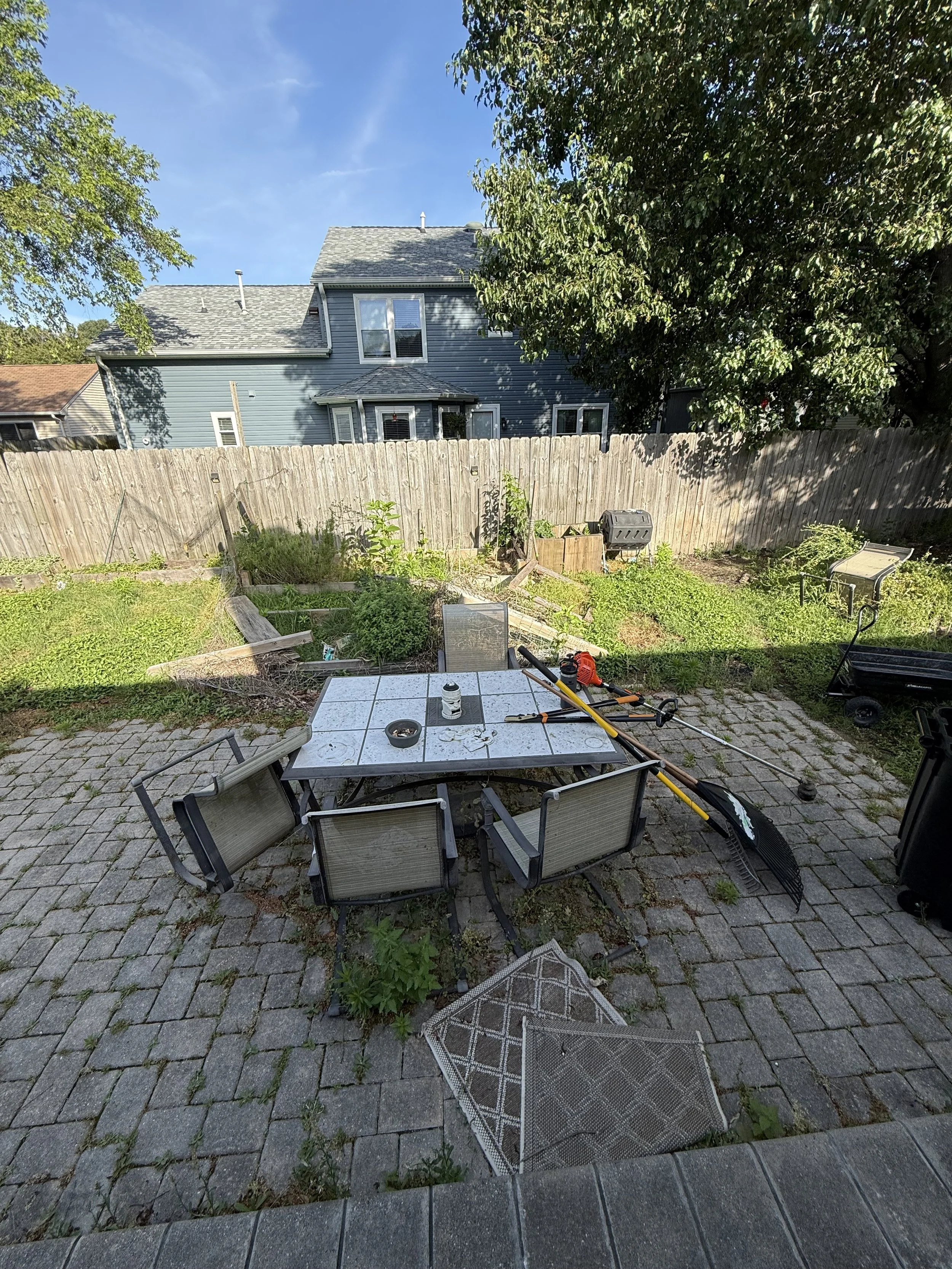 Backyard patio with a rectangular dining table, six chairs, and various yard tools and objects on the table. The patio is paved with bricks, and there are some plants and weeds along the edges. A wooden fence encloses the yard, with a blue house and 