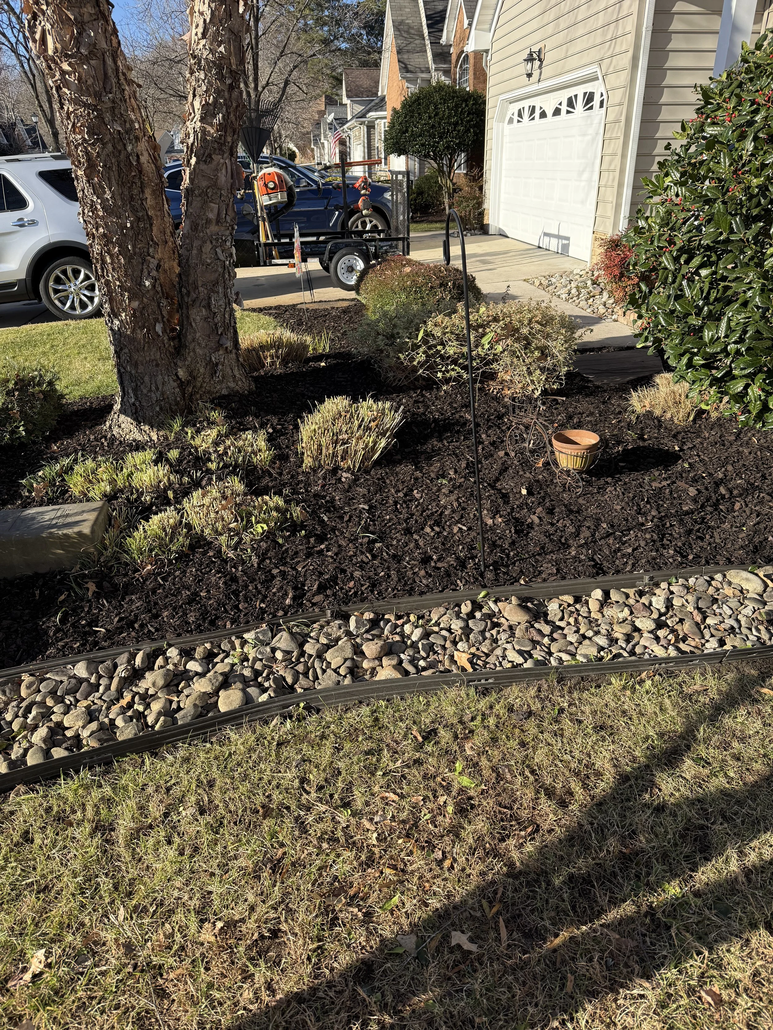 Front yard garden with two trees, shrubs, flowerpots, and a mulch bed with a border of small rocks. Houses and vehicles are visible in the background.