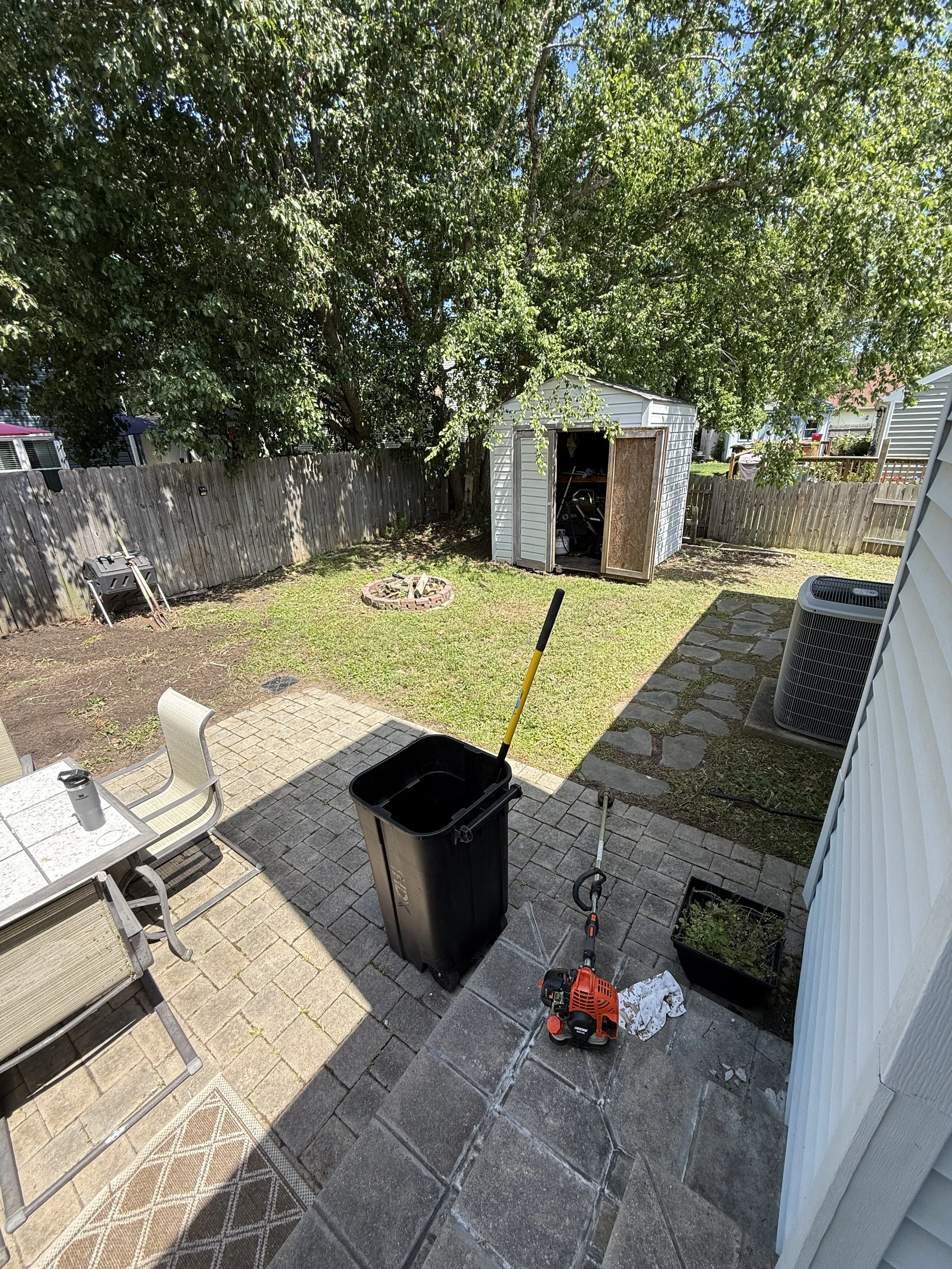 Backyard with patio, trash can, leaf blower, garden tools, and a shed near a wooden fence, surrounded by trees.