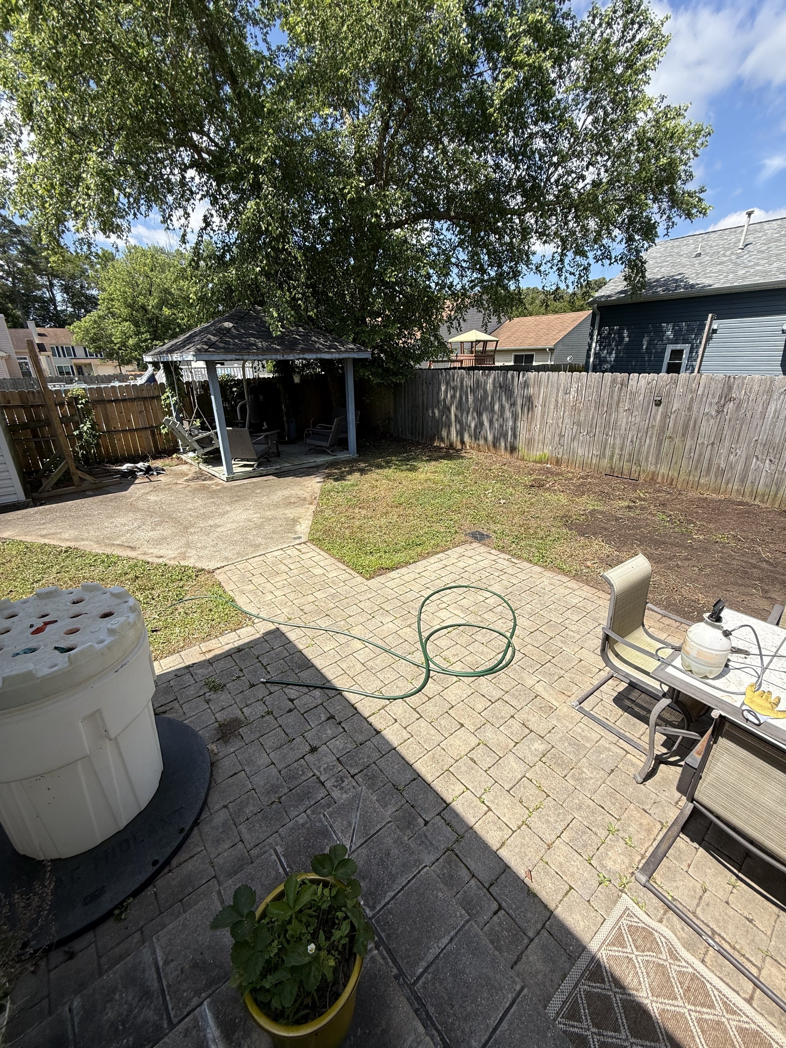 A backyard patio with a large tree providing shade, a gazebo with outdoor seating, a potted plant, a hose, and outdoor furniture on a paved surface, enclosed by a wooden fence.
