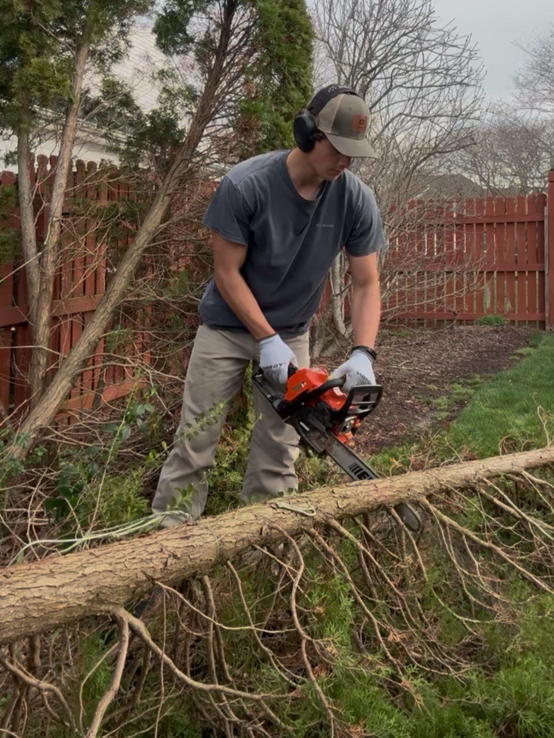 A young man wearing a baseball cap, black headphones, gray T-shirt, and gloves uses a leaf blower to clear fallen branches in a backyard with a wooden fence and trees.